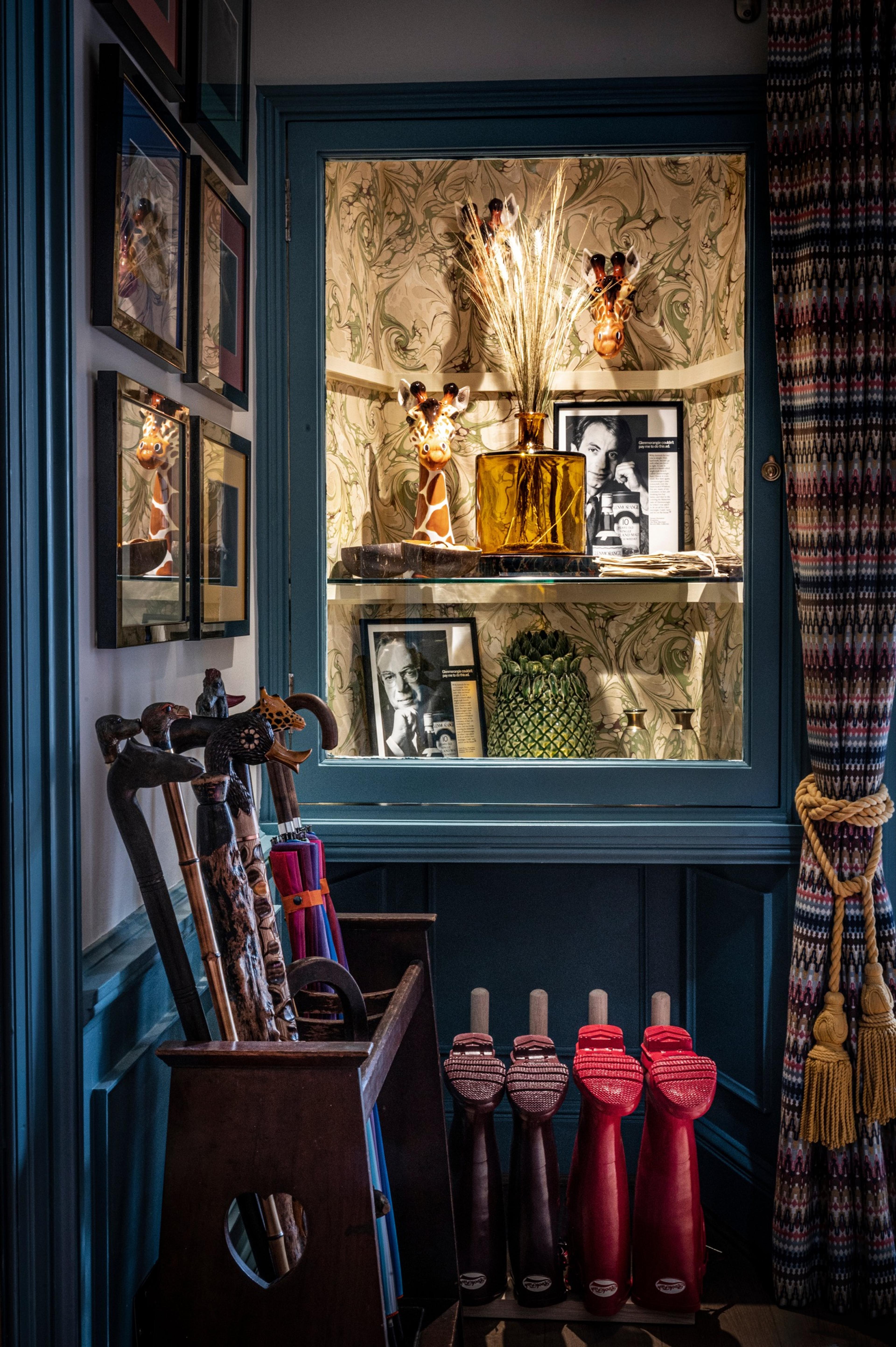 A corner of a room with a glass-fronted cabinet of ornaments and photographs, walking sticks in a stand and red wellington boots lined up below
