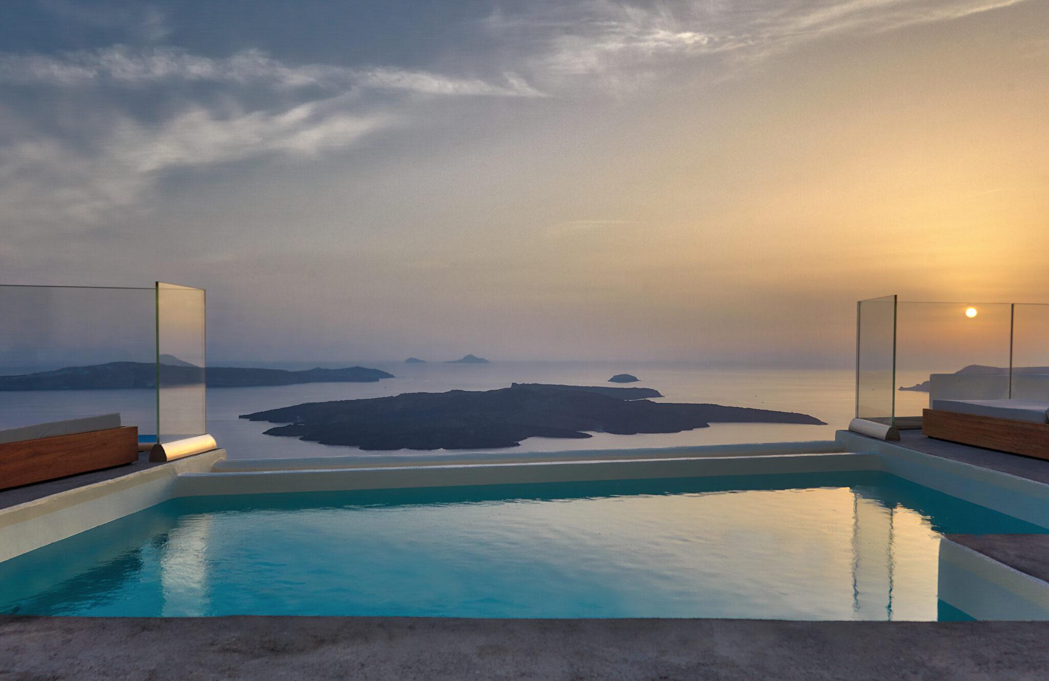 An infinity edge pool at sunset, reflecting an orange sky with small islands on the horizon.