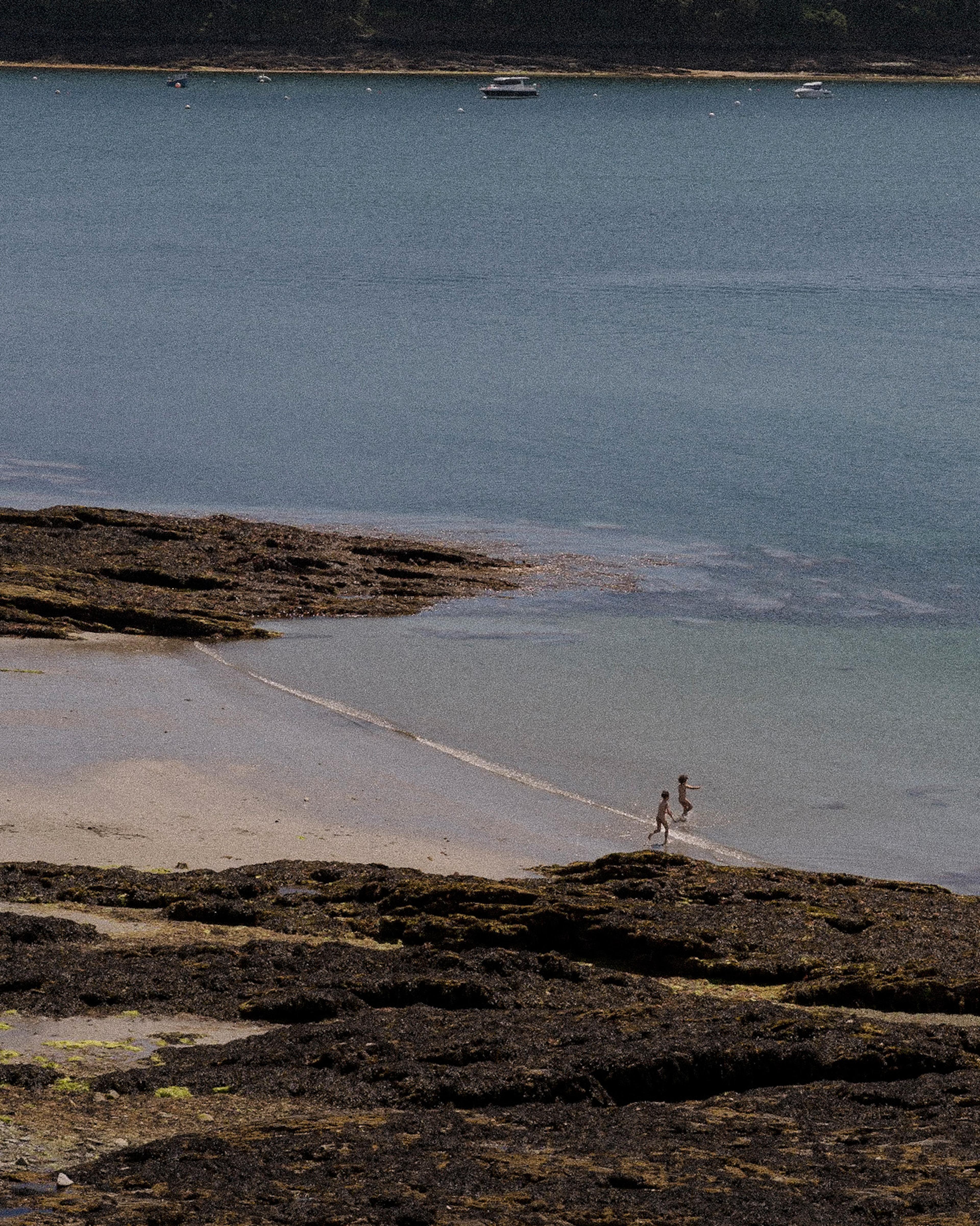 Two children running through shallow water at low tide, with boats anchored offshore.