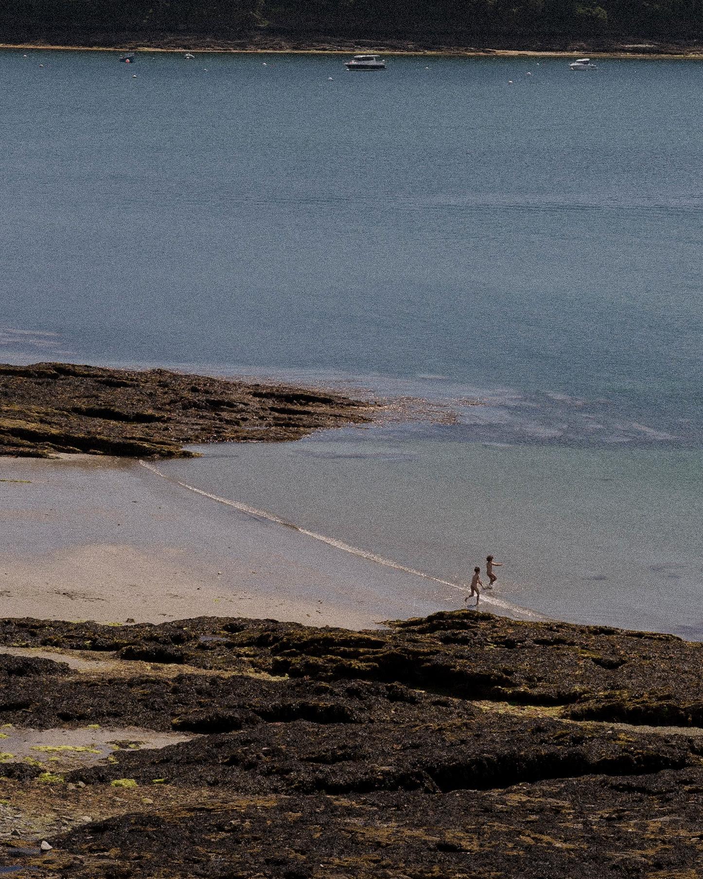 Two children running through shallow water at low tide, with boats anchored offshore.