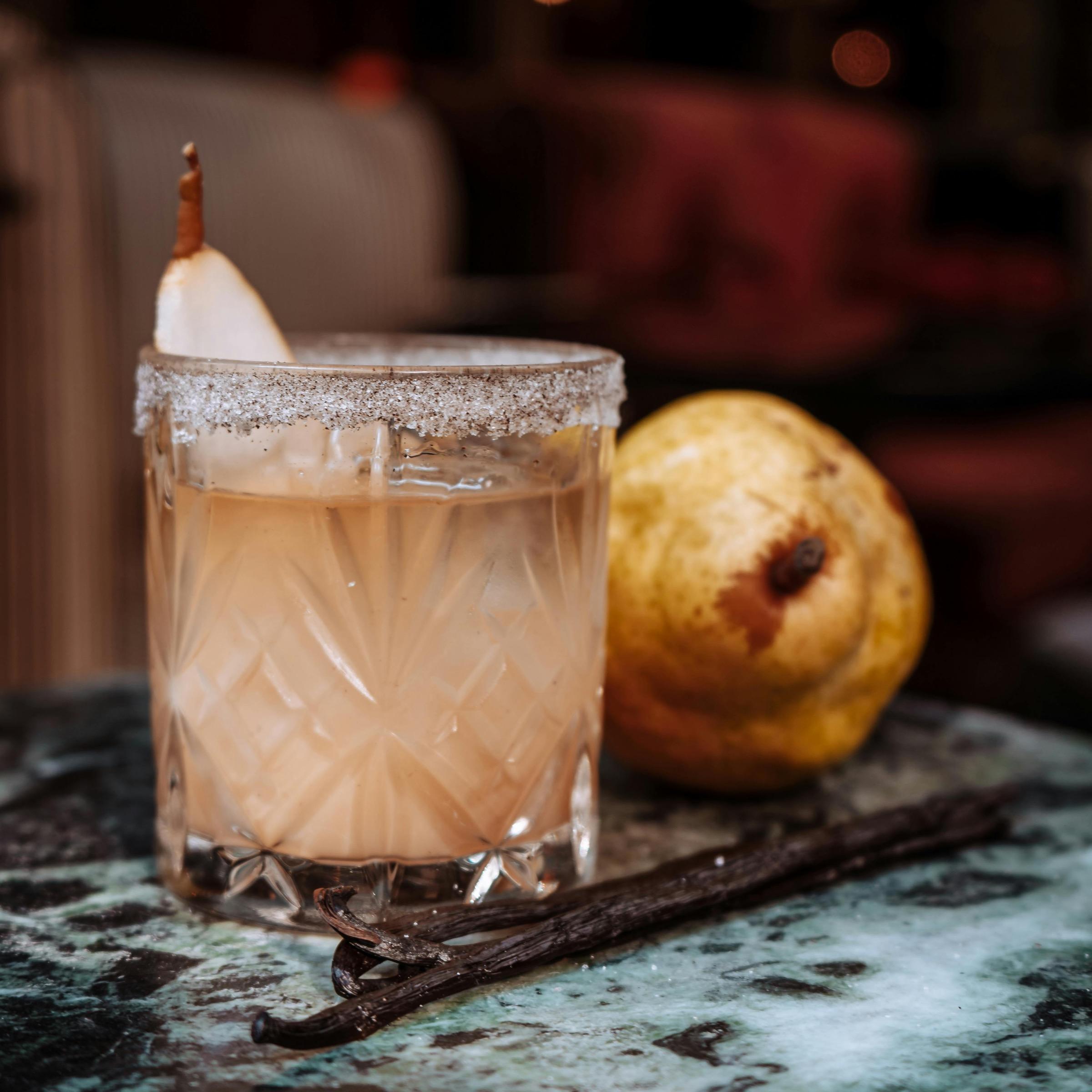 A sugar-rimmed cocktail garnished with a dried pear slice, set beside a whole pear and vanilla pods on a marble table.
