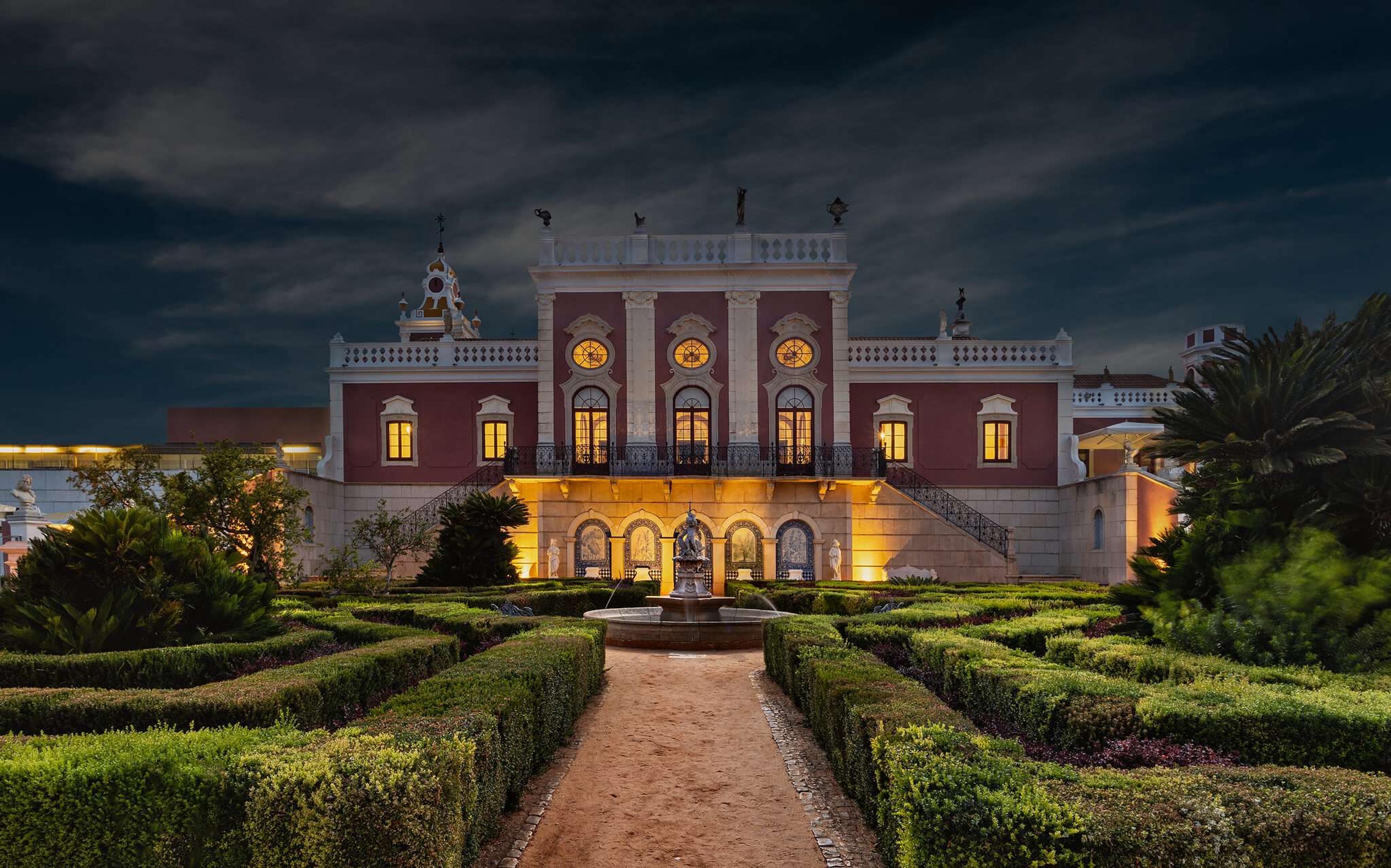 The palace at night, its rose-coloured façade lit from below, with a stone fountain and clipped hedges in the garden in front.