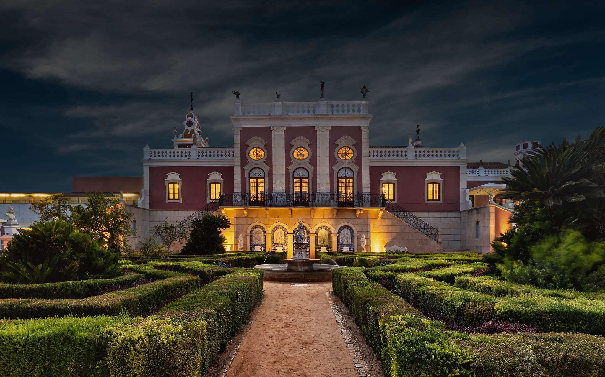 The palace at night, its rose-coloured façade lit from below, with a stone fountain and clipped hedges in the garden in front.