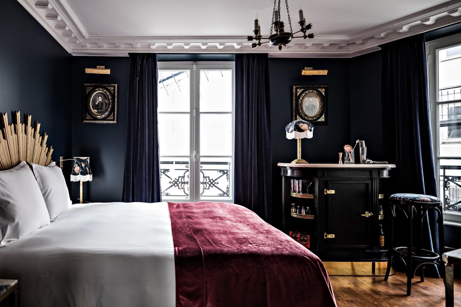 A bedroom with navy blue walls and curtains, a gold sunburst headboard, a burgundy velvet throw on the bed, a black cabinet, a stool, and tall French windows opening onto a wrought iron balcony.