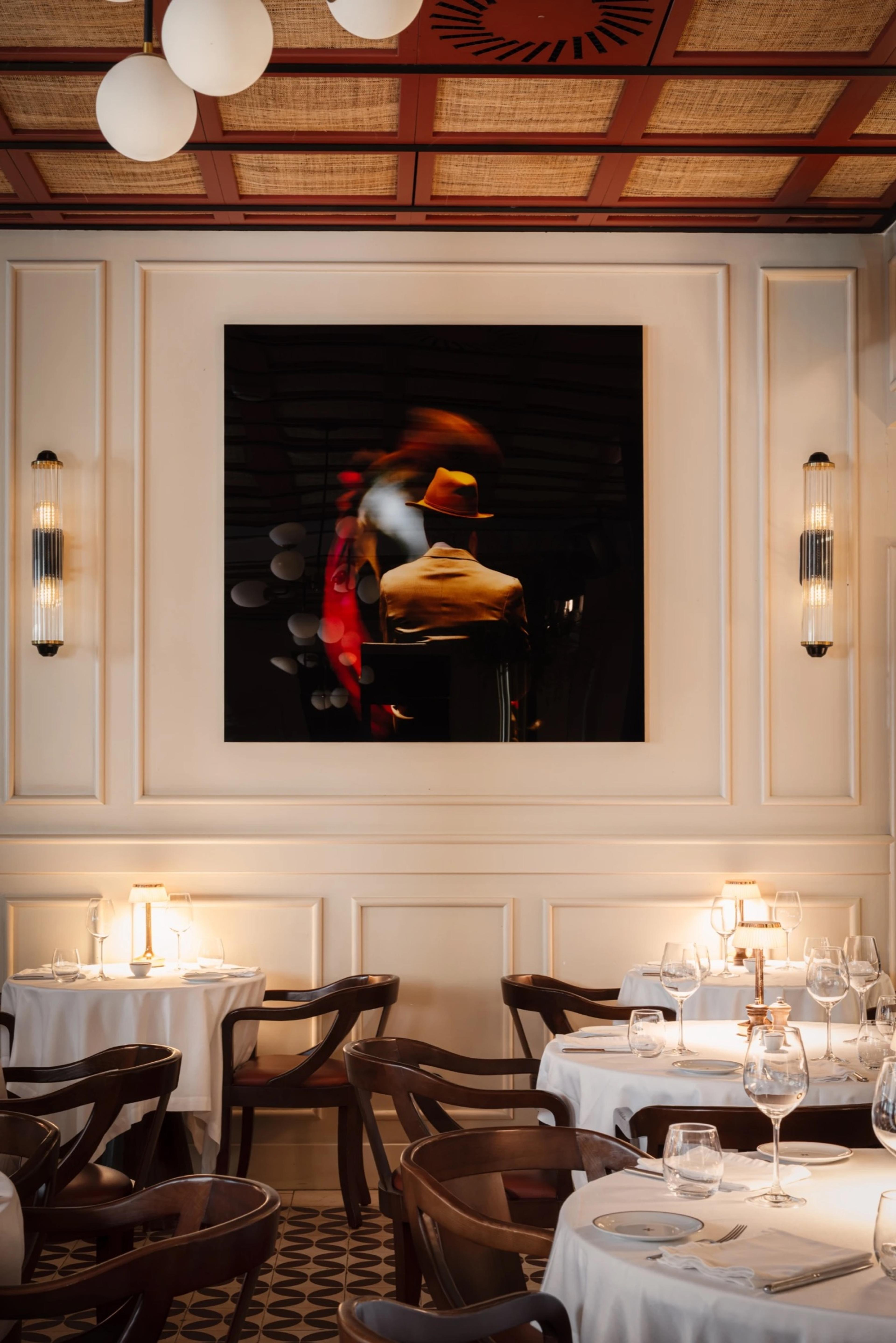 A dining room with cream panelled walls, a large dark framed photograph of a man in a hat, white-clothed tables and dark wooden bentwood chairs, under a rattan ceiling.