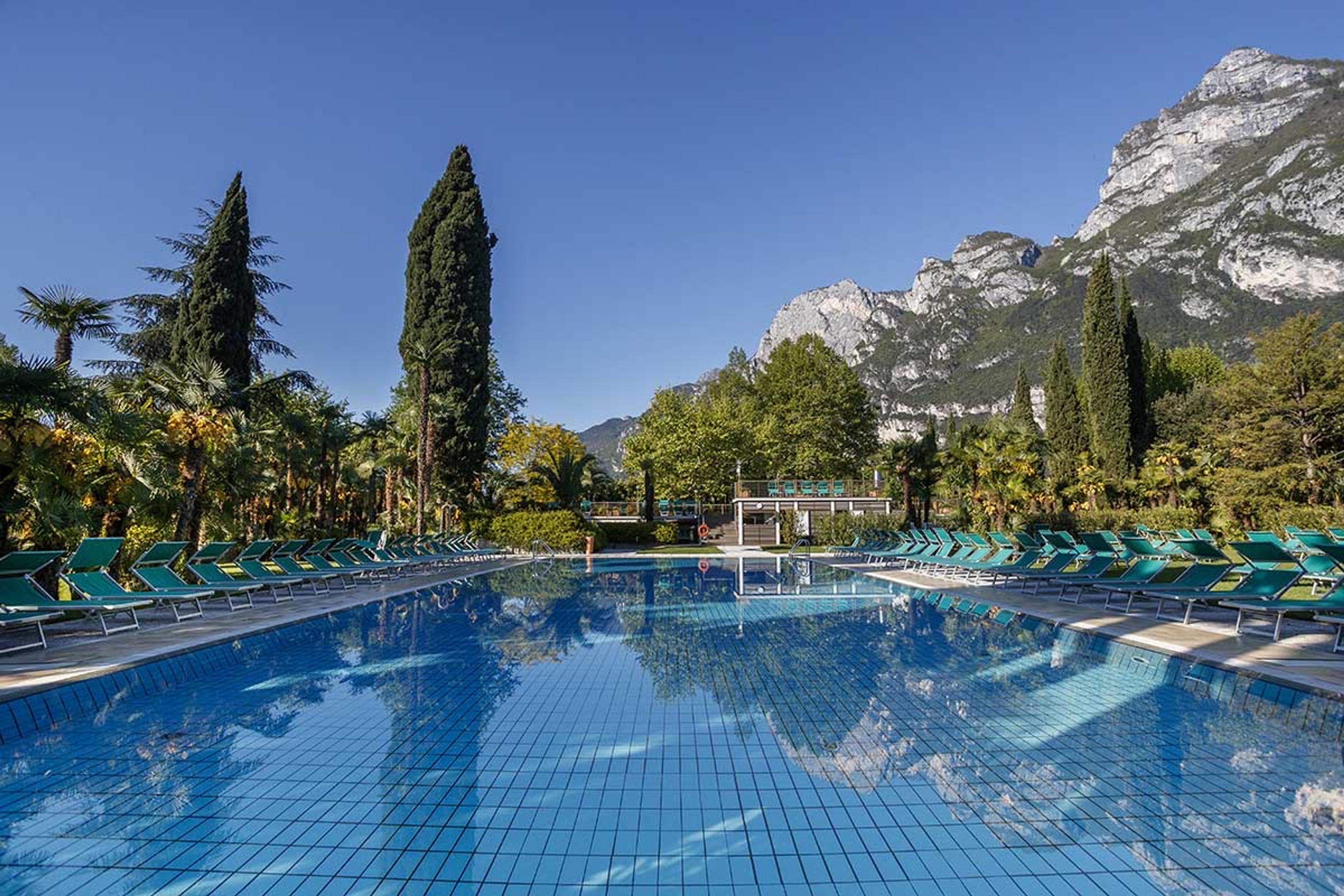 An outdoor swimming pool with turquoise tiled edges and green loungers along its sides, with cypress and palm trees beyond, a rocky mountain behind, and nobody in it.