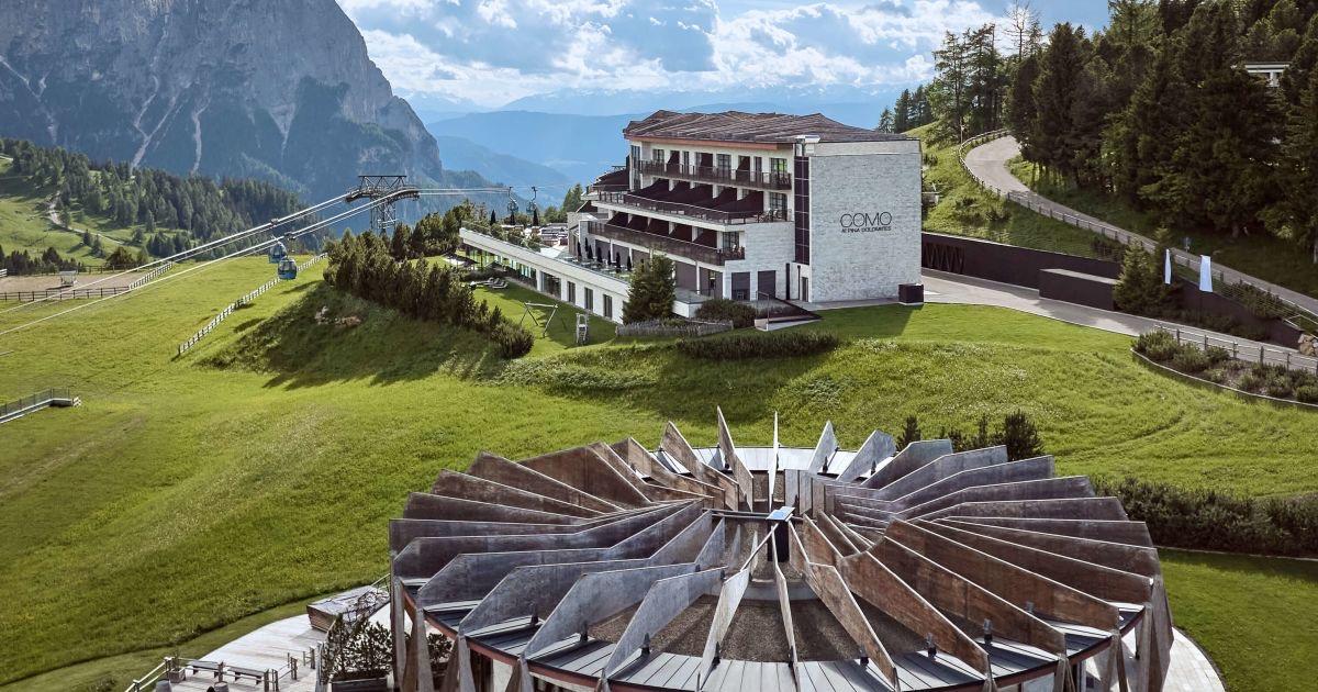 A hotel building and a circular open-air restaurant with a fanned timber roof sit on a grassy hillside below a jagged grey peak, with a cable car line crossing the slope behind them.