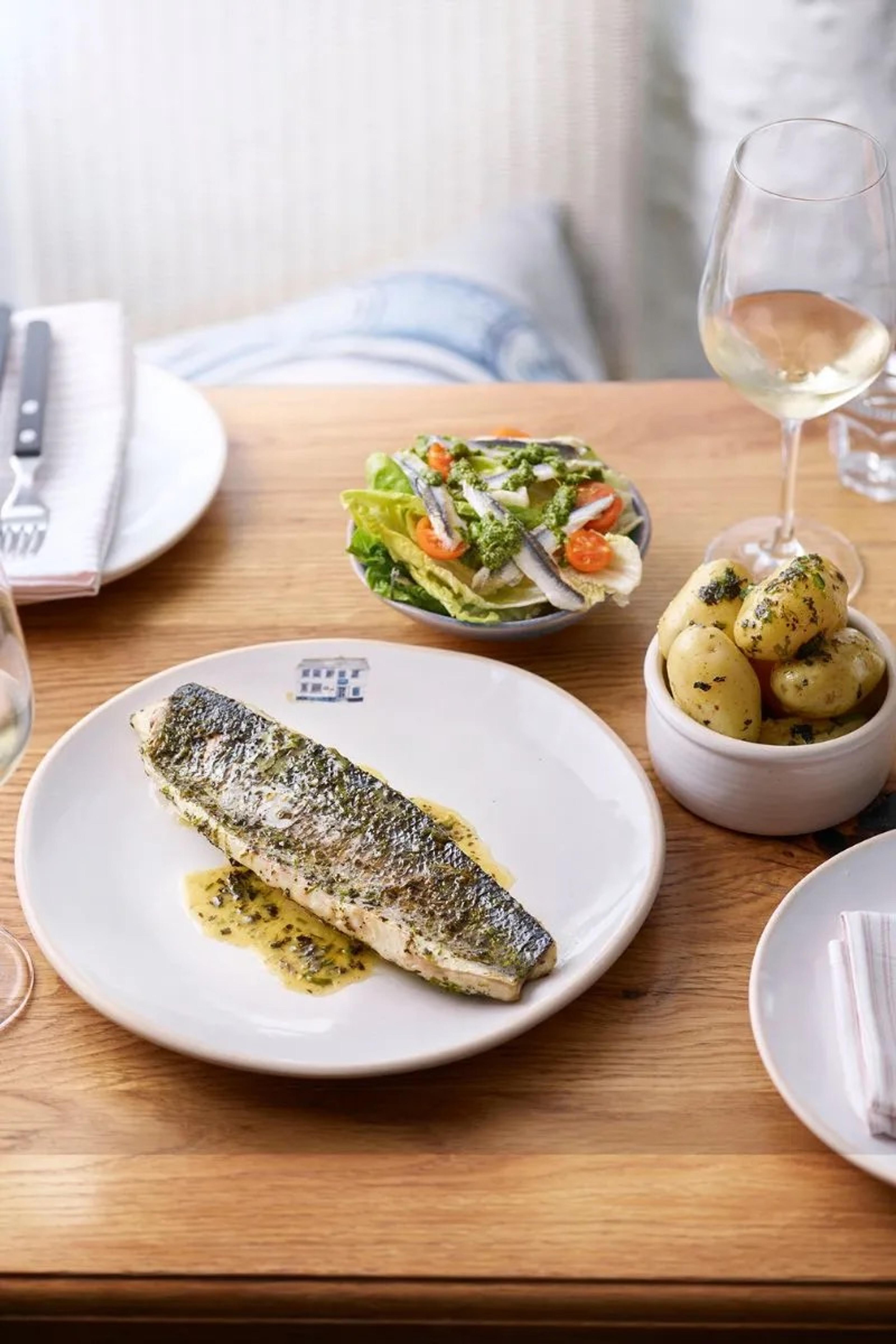 A grilled fish fillet in butter sauce, with a bowl of salad and a dish of new potatoes beside it.