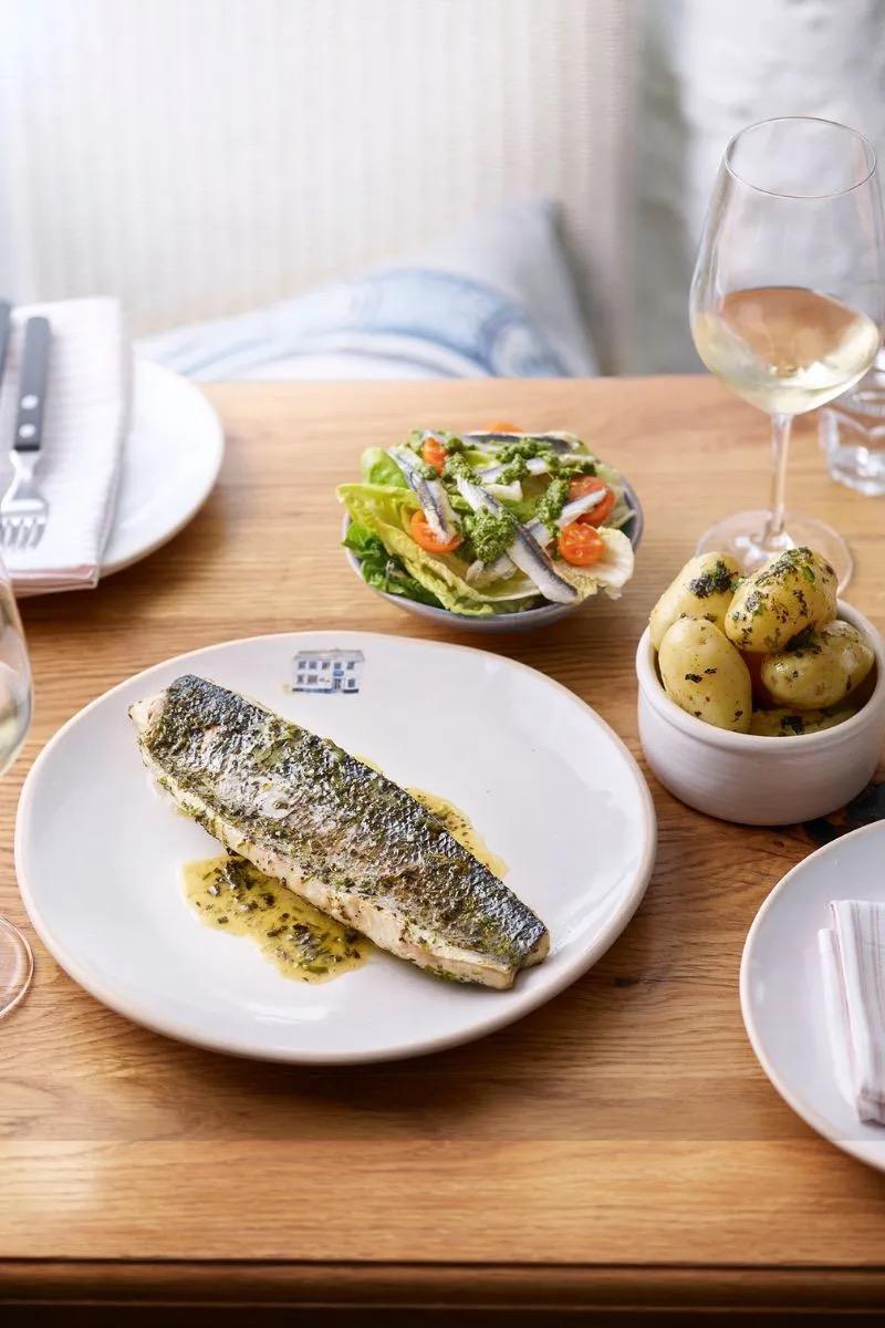 A grilled fish fillet in butter sauce, with a bowl of salad and a dish of new potatoes beside it.