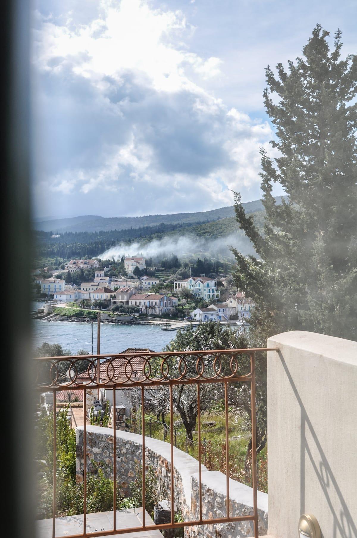 A view across a harbour town of white and pastel houses to wooded hills, seen past a wrought-iron balcony railing and a pine tree, with the dark edge of a window frame at the left.