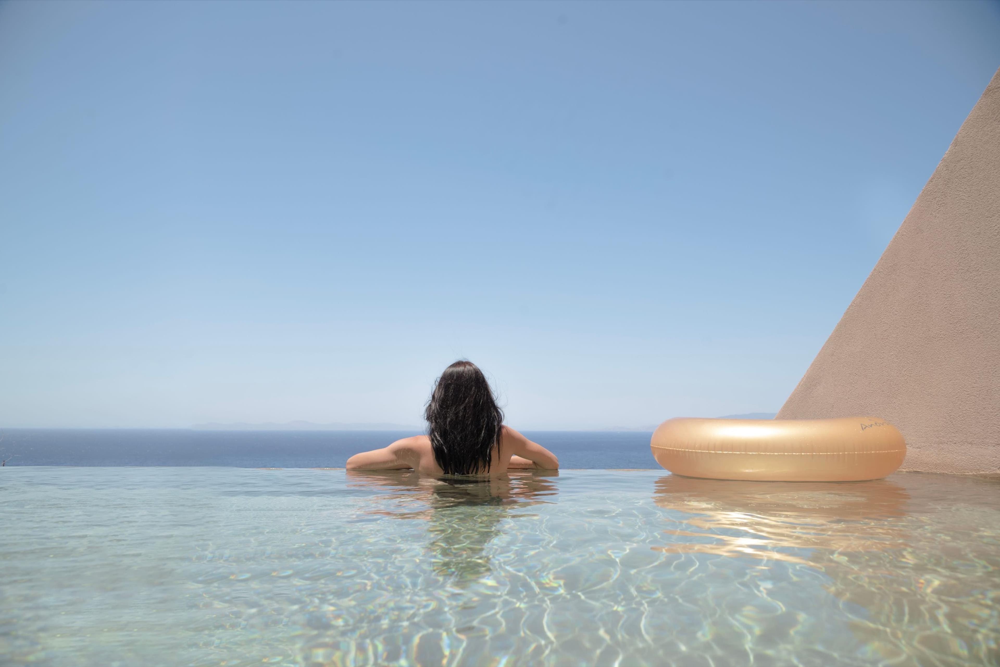 A woman floats in an infinity pool with her back to the camera, her wet hair visible above the waterline, beside an inflatable ring and a plain stone wall, with the sea on the horizon.