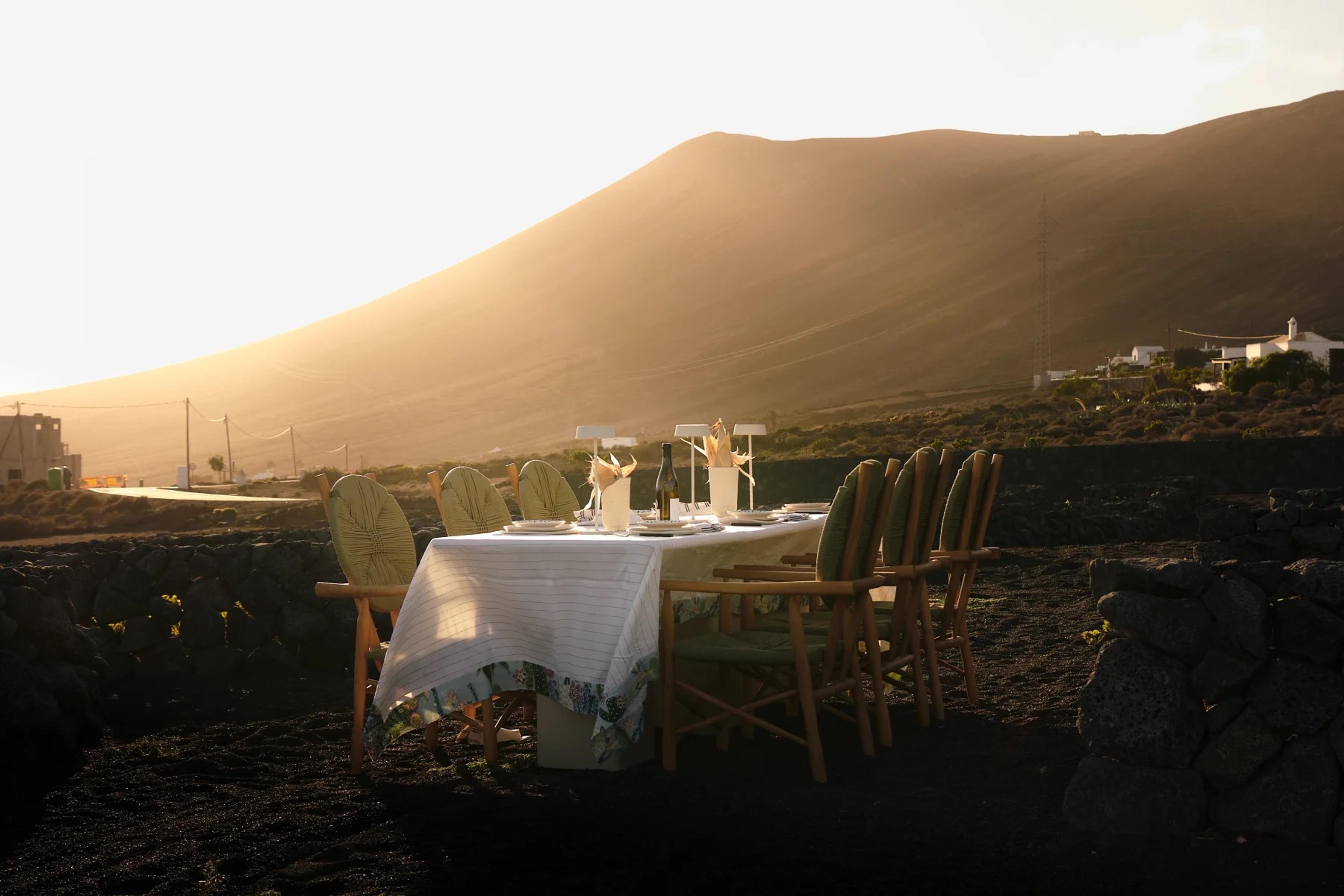 A long outdoor dining table set with white cloths, wine glasses and place settings on black volcanic ground, facing a bare mountain lit orange by the setting sun.