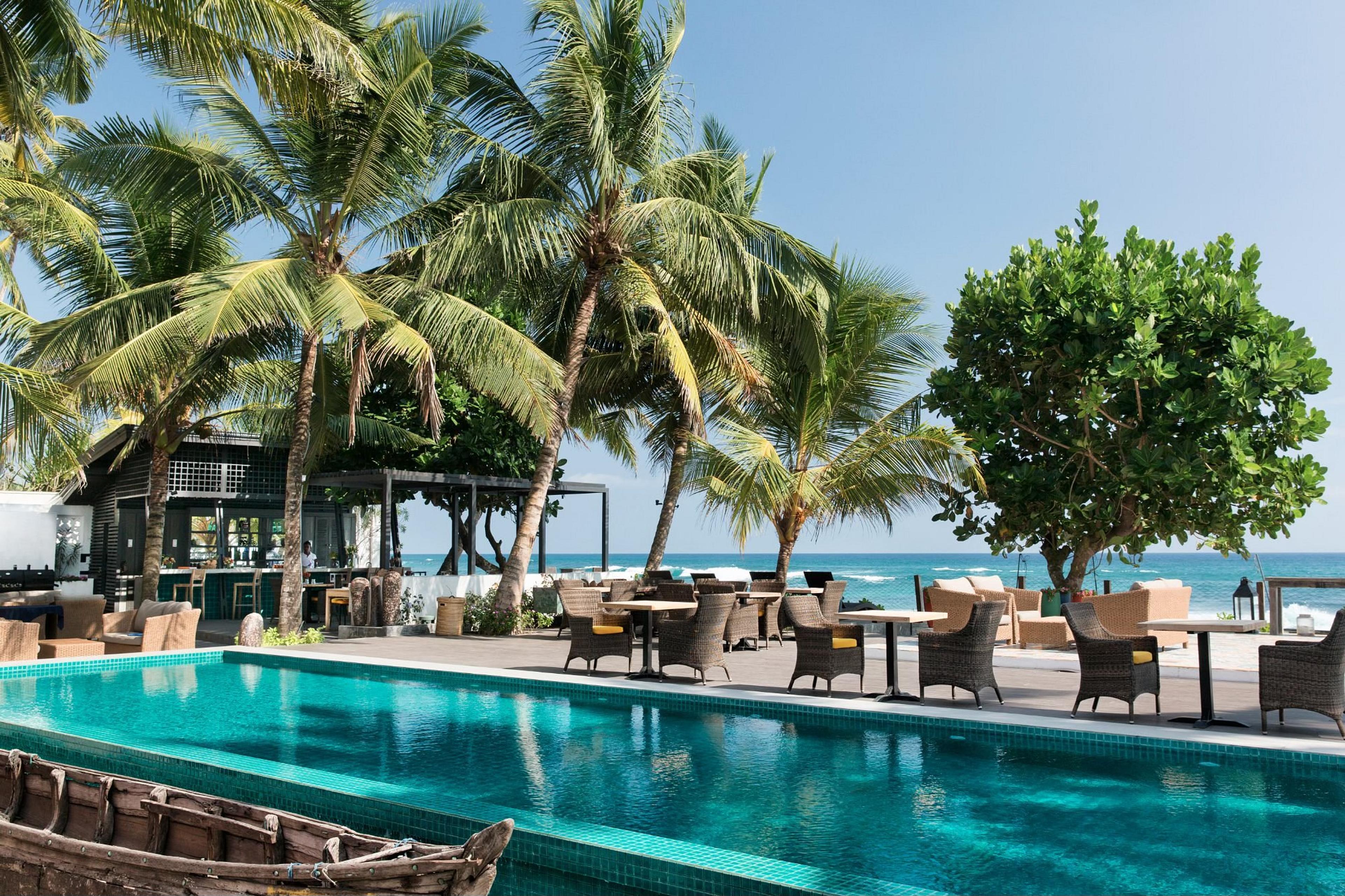 A turquoise pool beside a deck of wicker chairs and tables under coconut palms, a bar pavilion to one side and the Indian Ocean behind, with an old wooden boat in the foreground