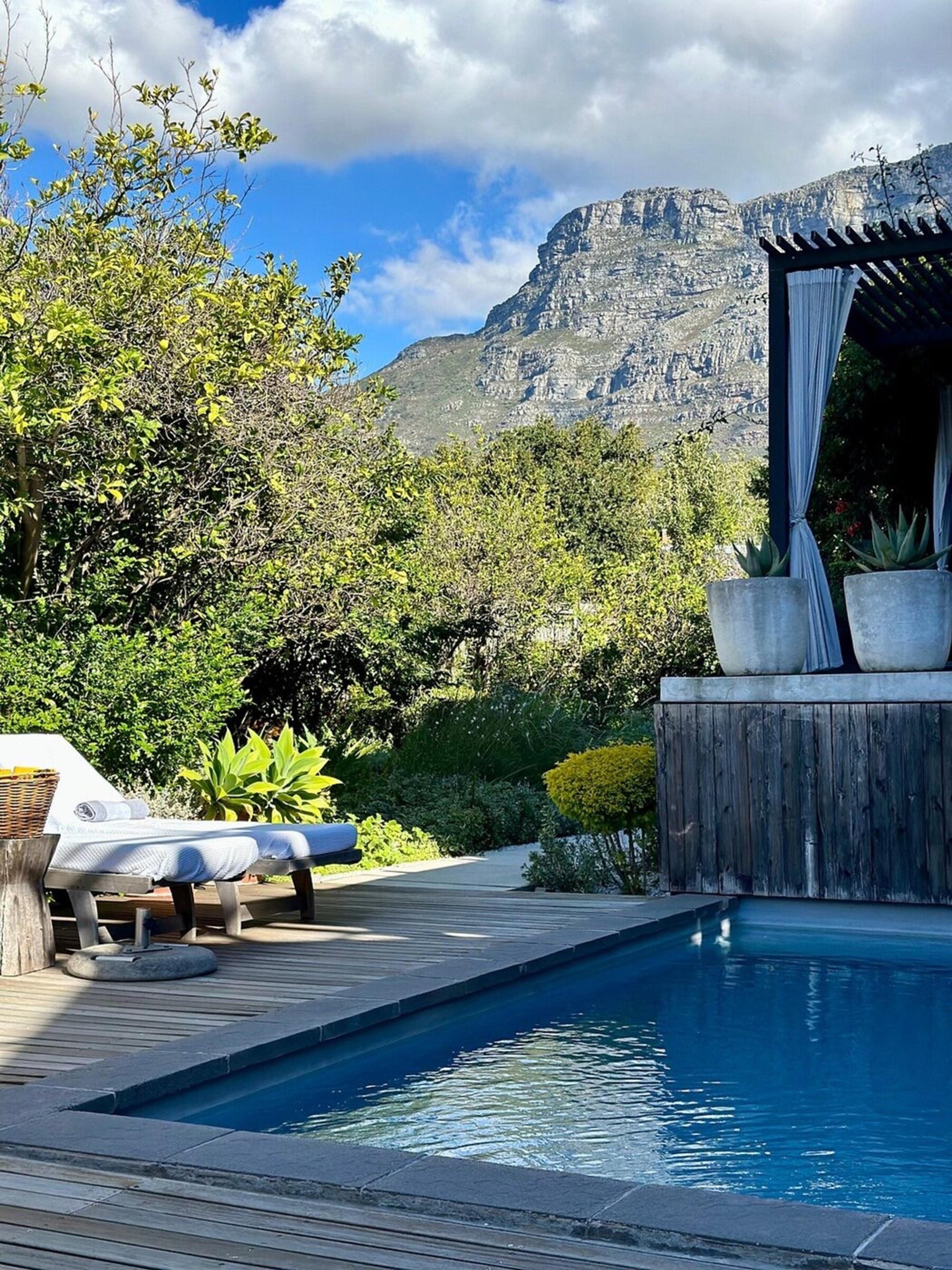 A rectangular pool beside a timber deck, with a white lounger, a dark pergola with pale curtains, and a rocky mountain above the trees.