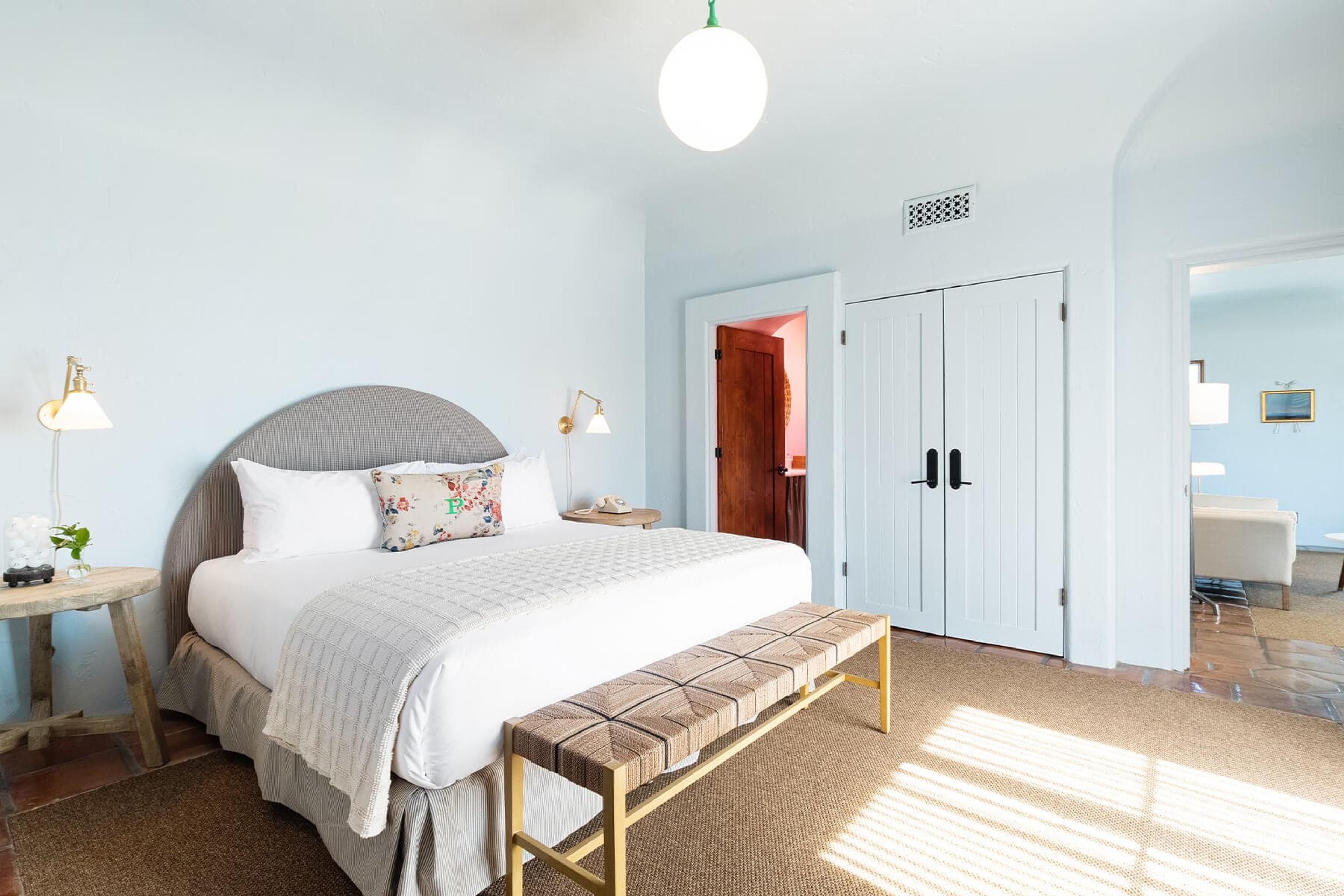 A made bed with a grey upholstered headboard and a woven bench at its foot, in a bright room with white louvre doors and a wood-topped side table.