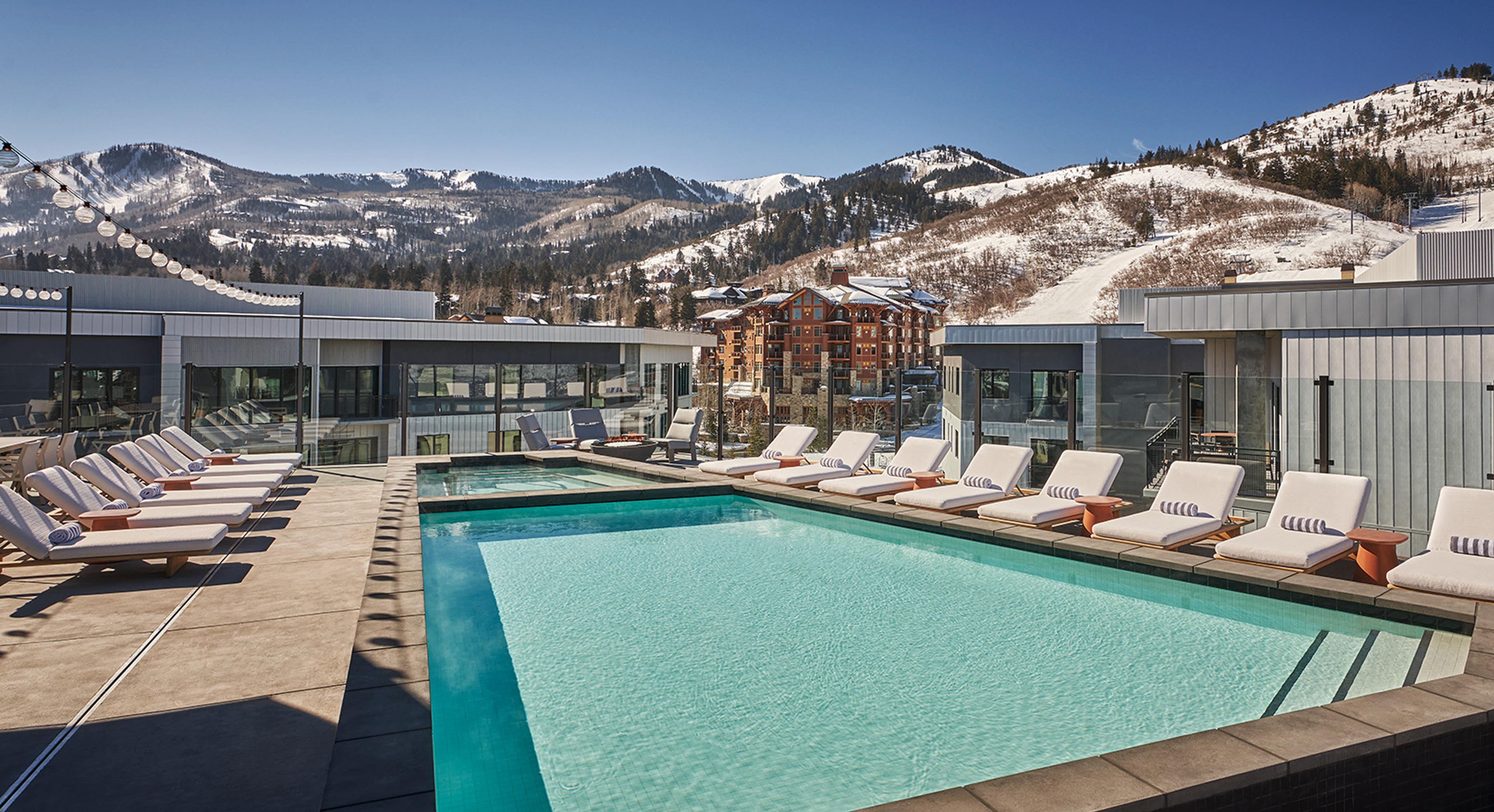 A rectangular rooftop pool of pale blue water, with a row of white loungers along its edge, a smaller pool beyond, and snowy mountains and buildings under a clear blue sky.