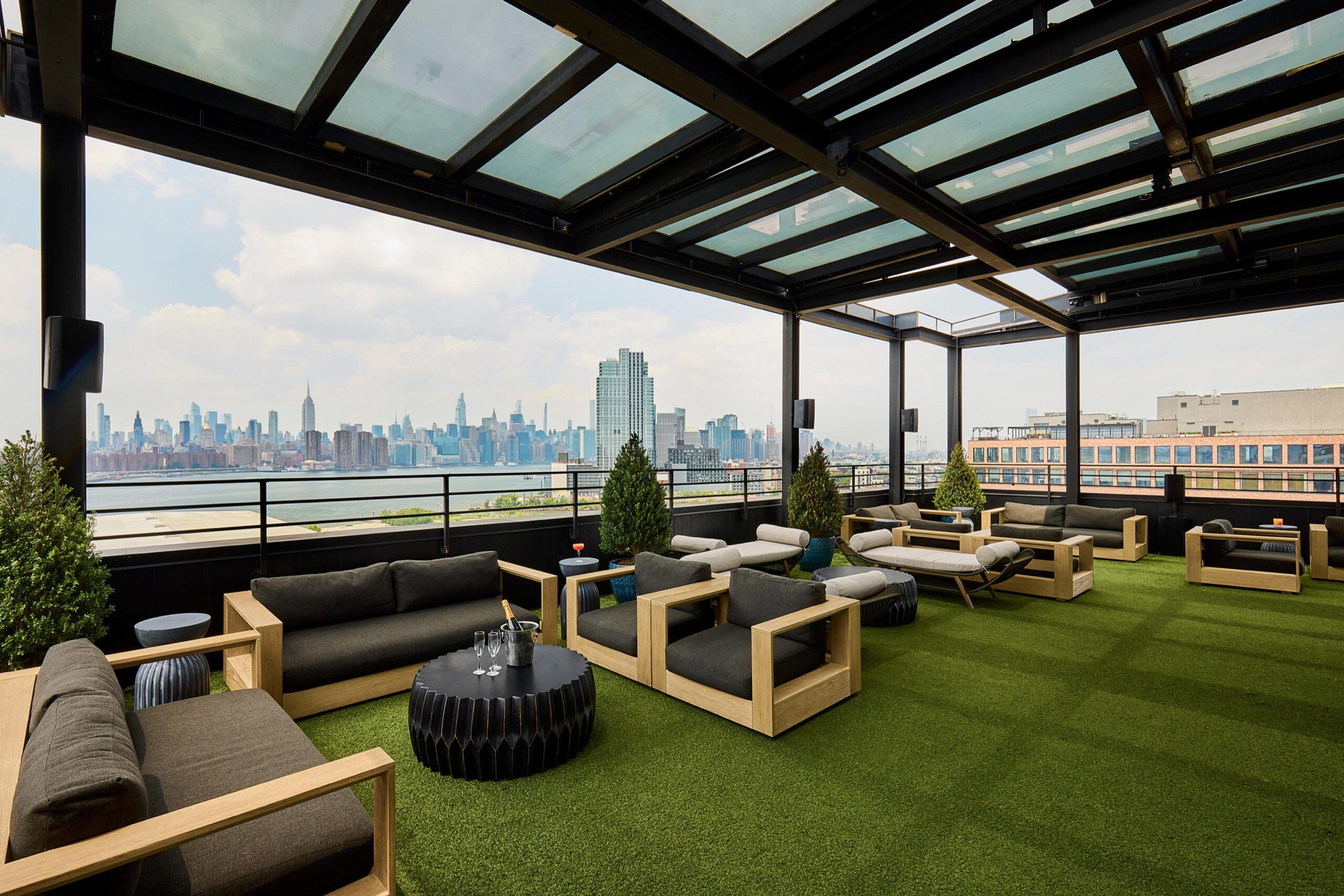 A covered rooftop terrace with wooden-framed sofas and armchairs on artificial turf, looking out over a river to the Manhattan skyline.