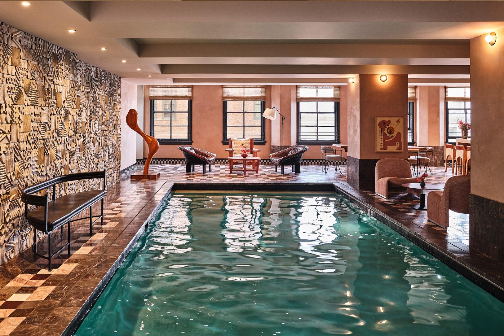 An indoor swimming pool with a carved wood sculpture and leather lounge chairs beside a textured stone wall, with a seating area and dining tables beyond.