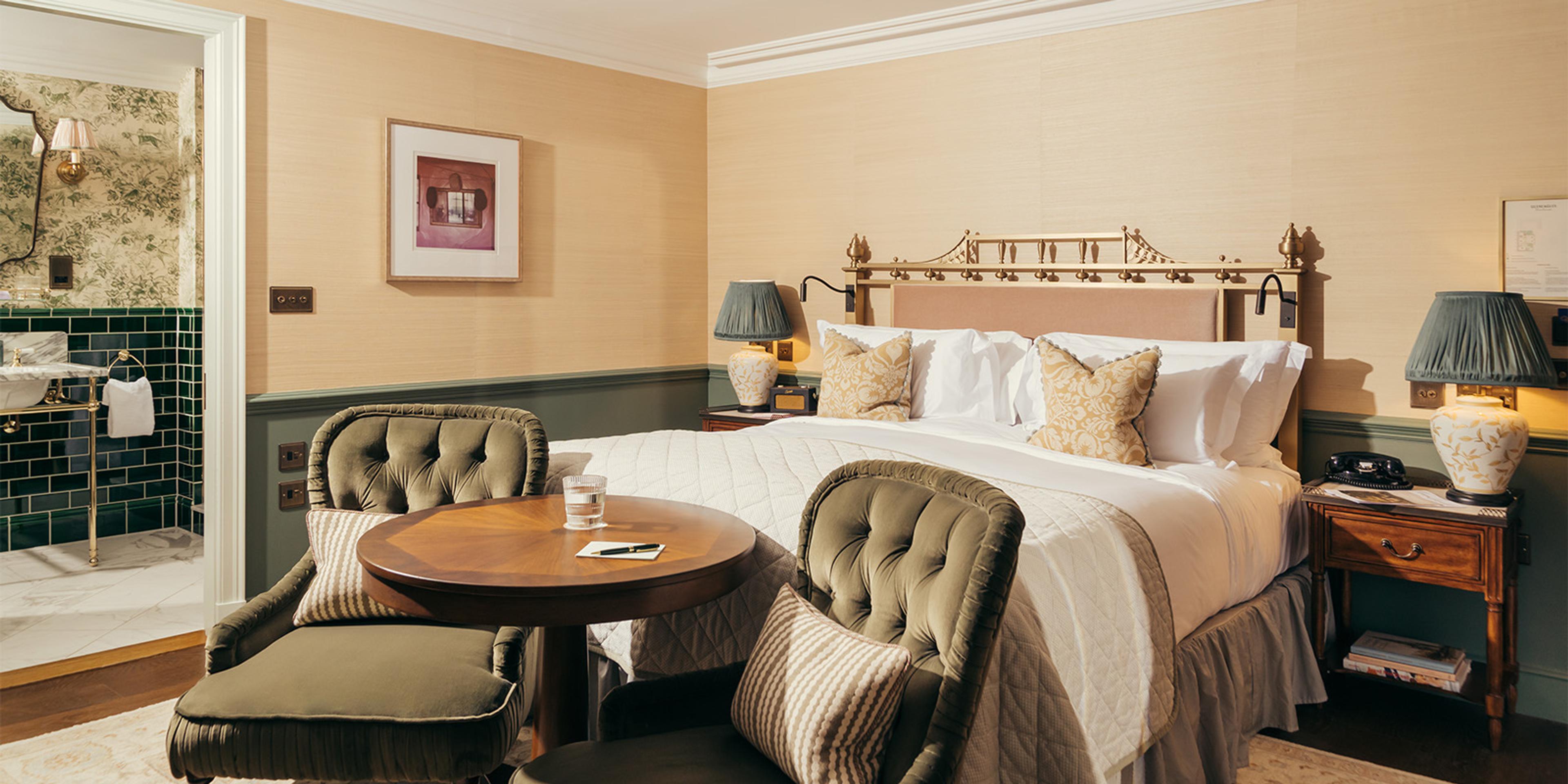 a bedroom with a pale bed, two tufted olive armchairs around a round table, and an open door to a green-tiled bathroom beyond
