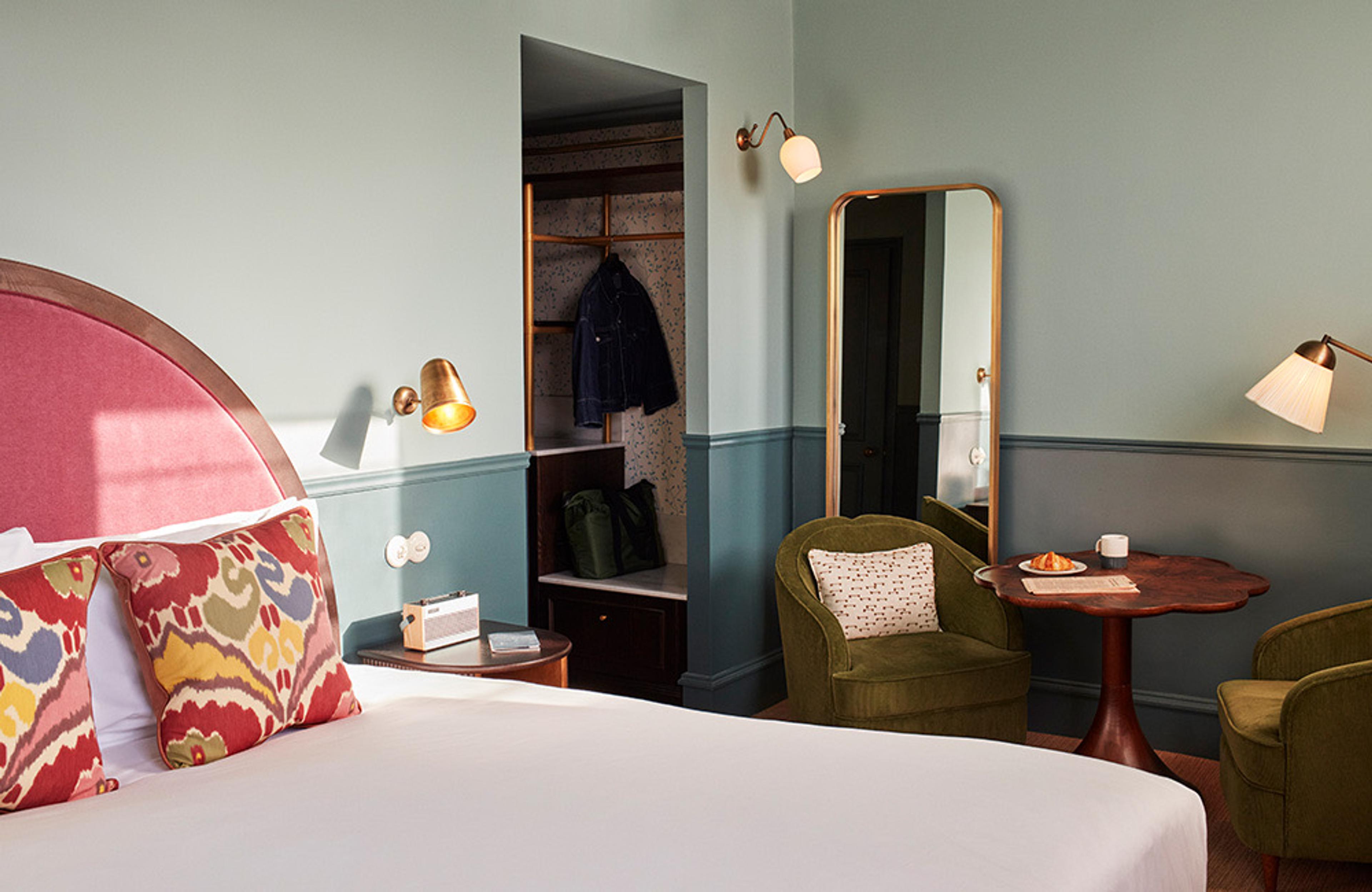 A bedroom with a red headboard and patterned cushions, a round table and armchair by the window and a mirror on the far wall.