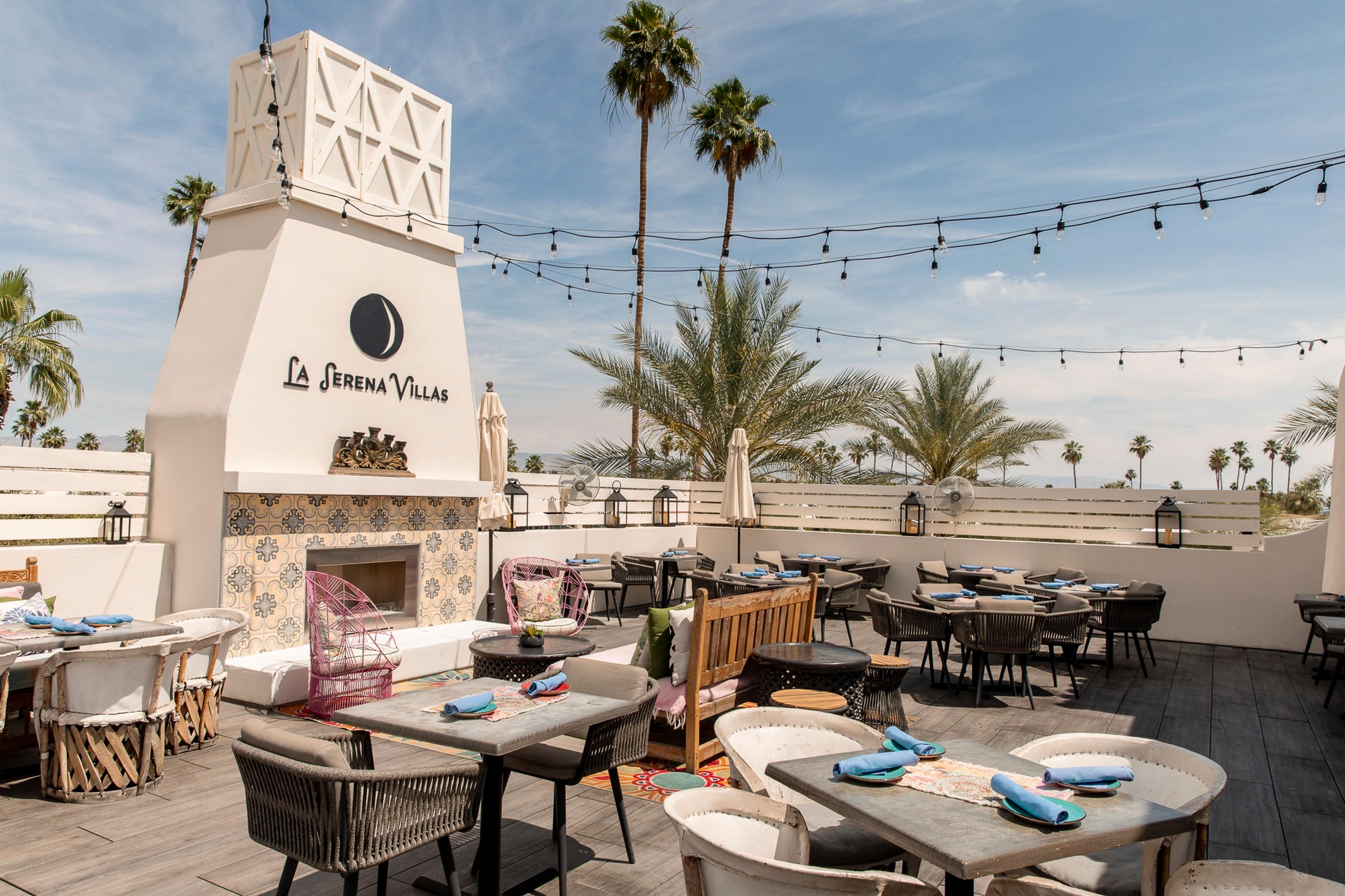 A rooftop terrace with dining tables, a tiled fireplace carrying the La Serena Villas sign, string lights and palm trees against a blue sky.
