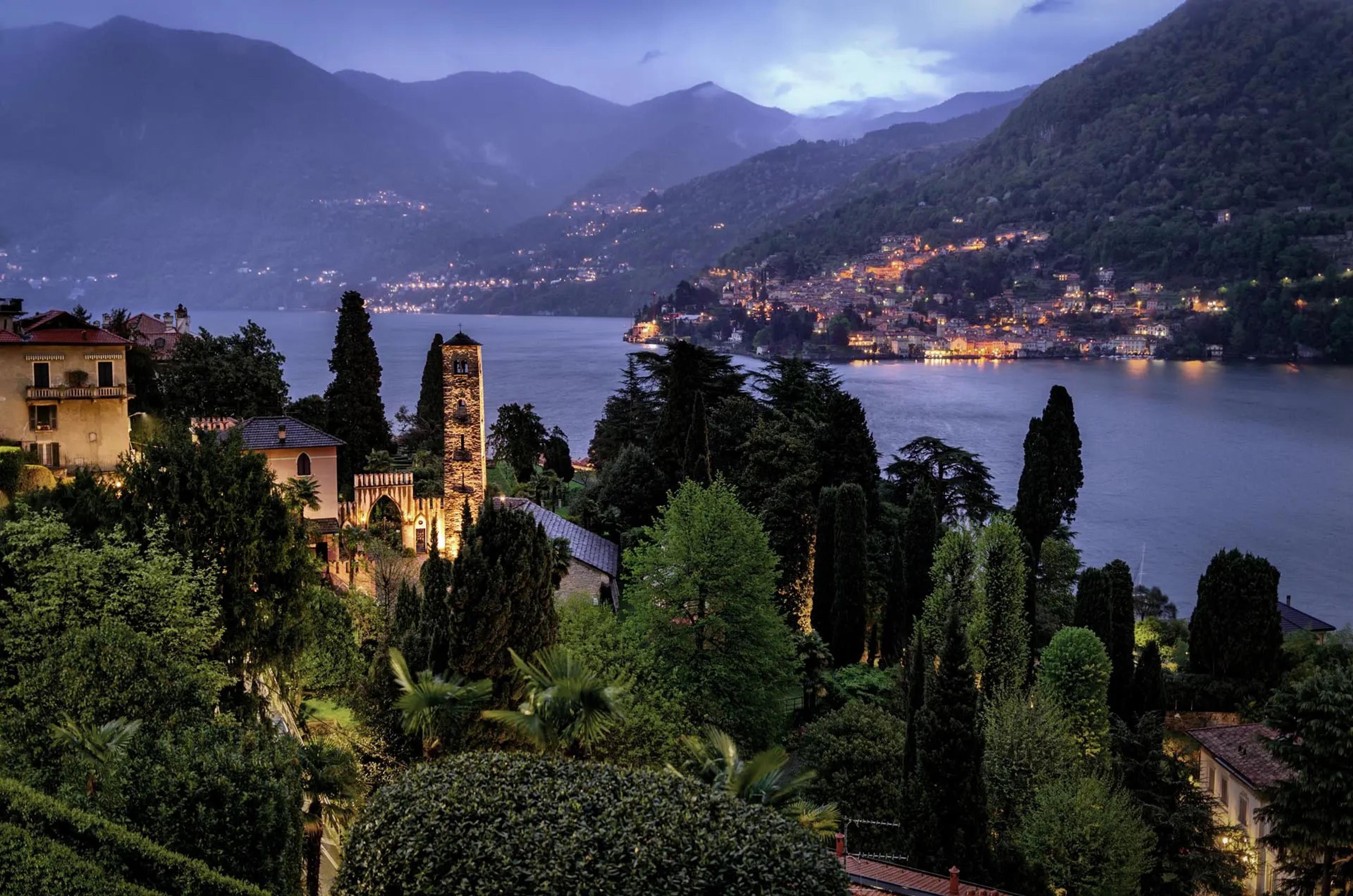 A lakeside town lit at dusk, seen past a stone bell tower and trees, with mountains rising beyond the water.
