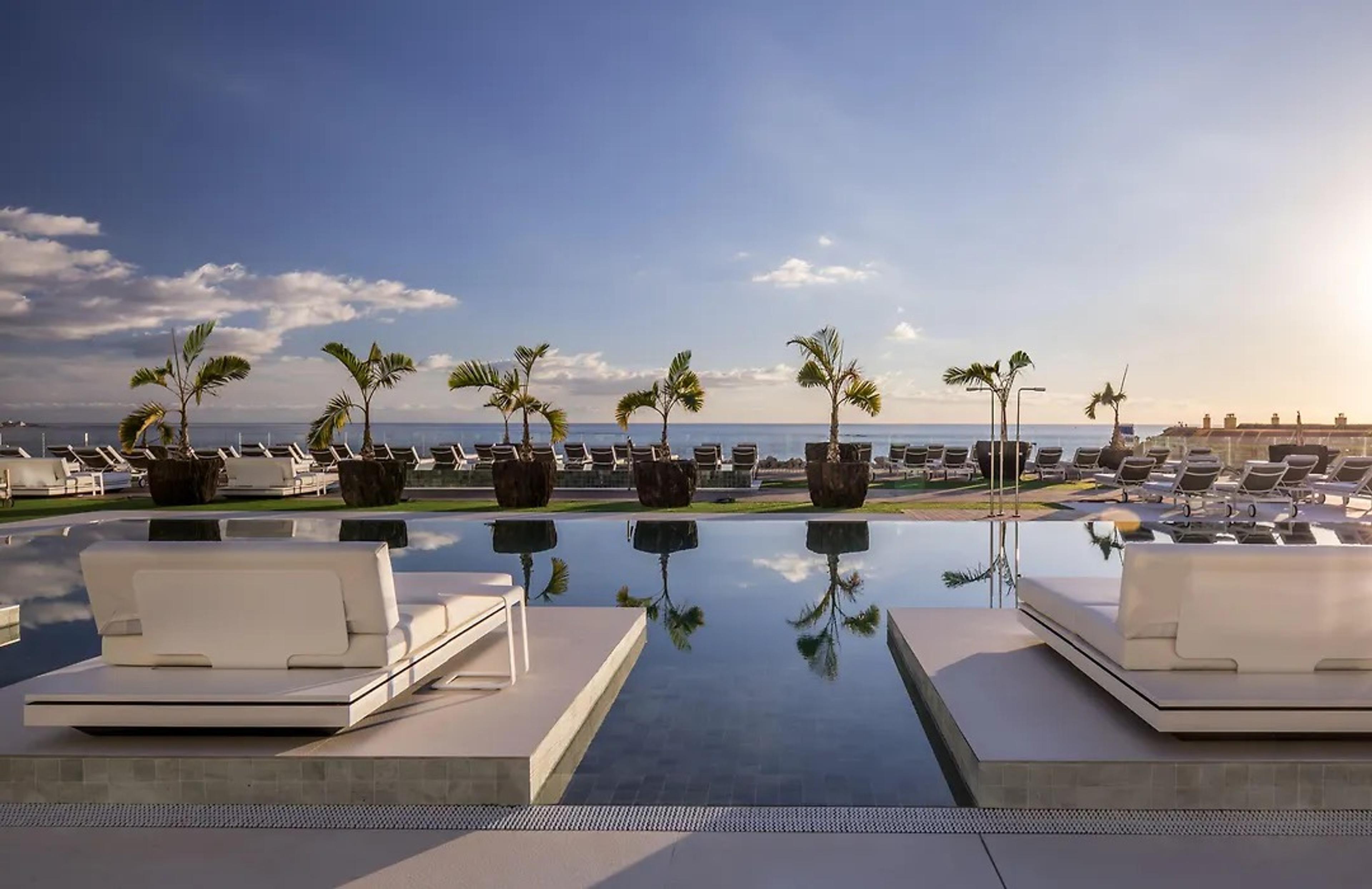 White daybeds on the edge of a still pool at sunset, potted palms reflected in the water and the sea behind
