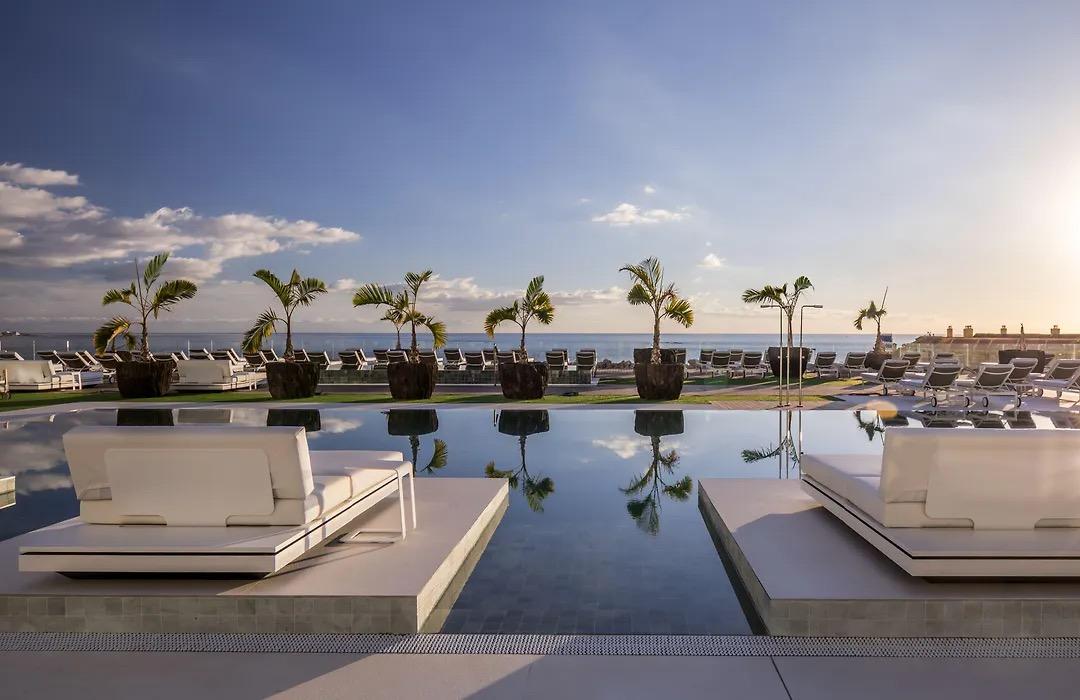 White daybeds on the edge of a still pool at sunset, potted palms reflected in the water and the sea behind