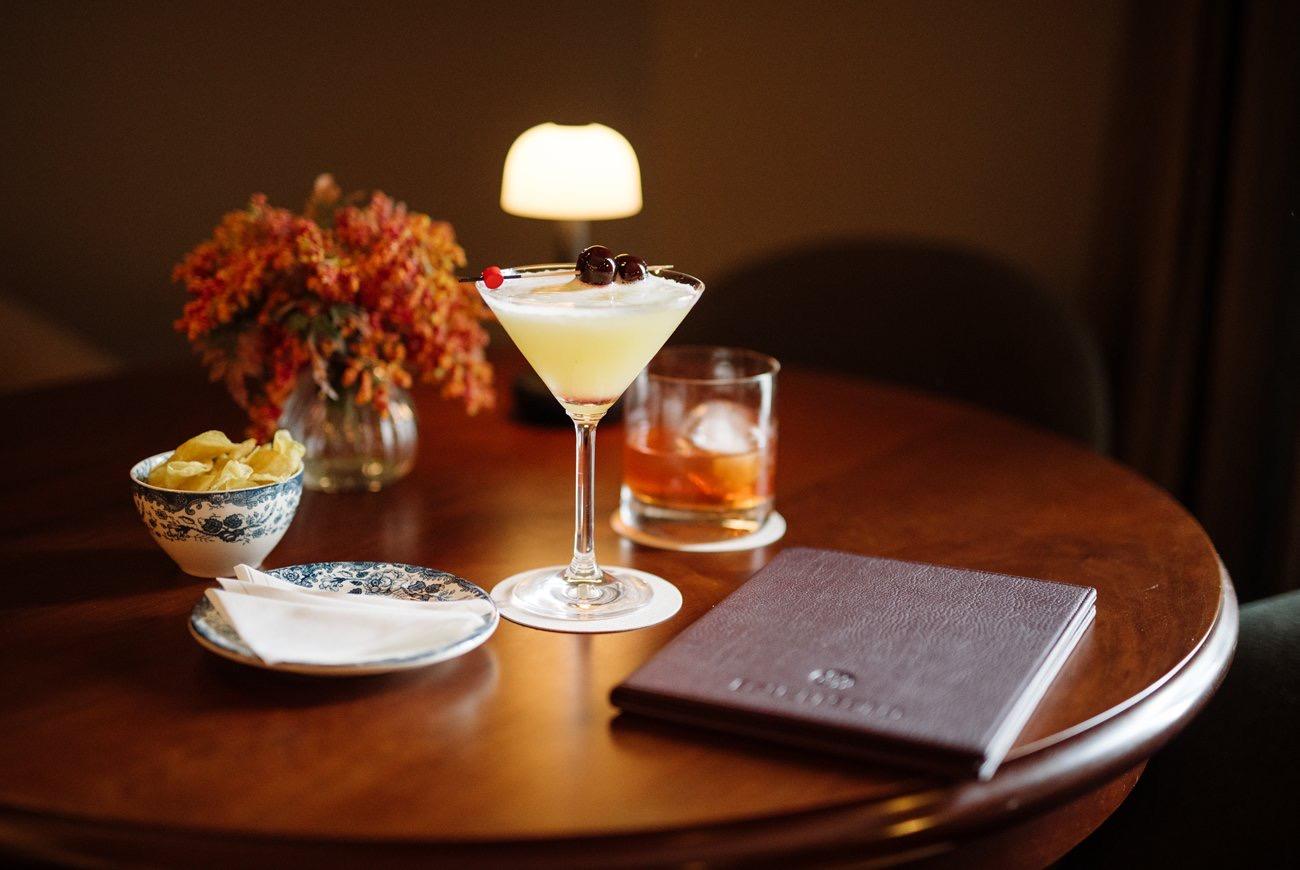A round wooden table holding a cocktail with cherries, a glass of whisky on ice, a bowl of crisps, a small dish, and a vase of orange flowers.