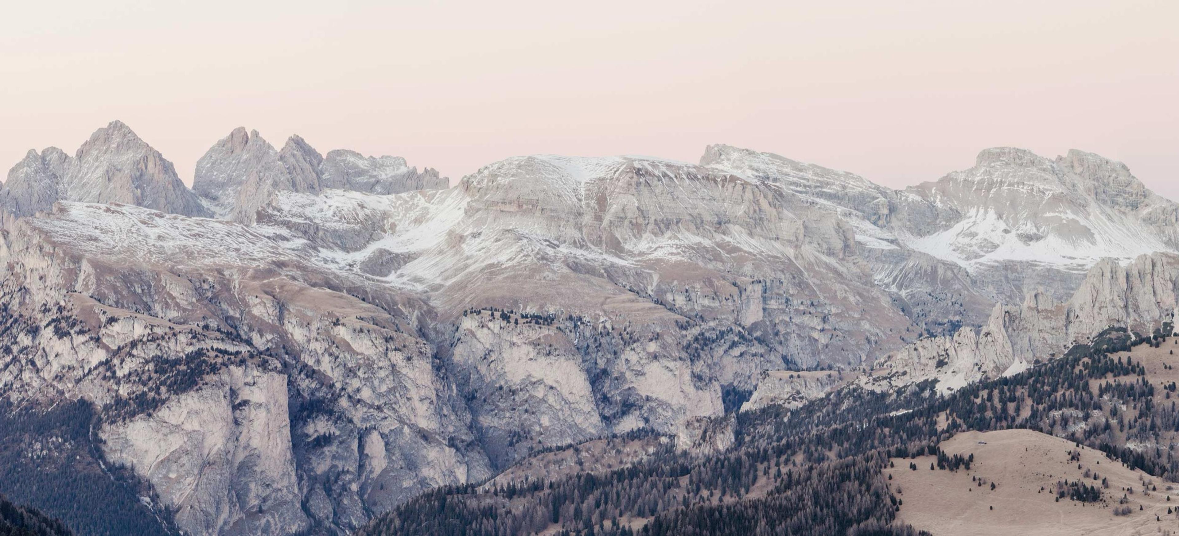 A panoramic view of the Dolomites mountain range at dusk, bare rock and snow patches above tree-covered lower slopes.