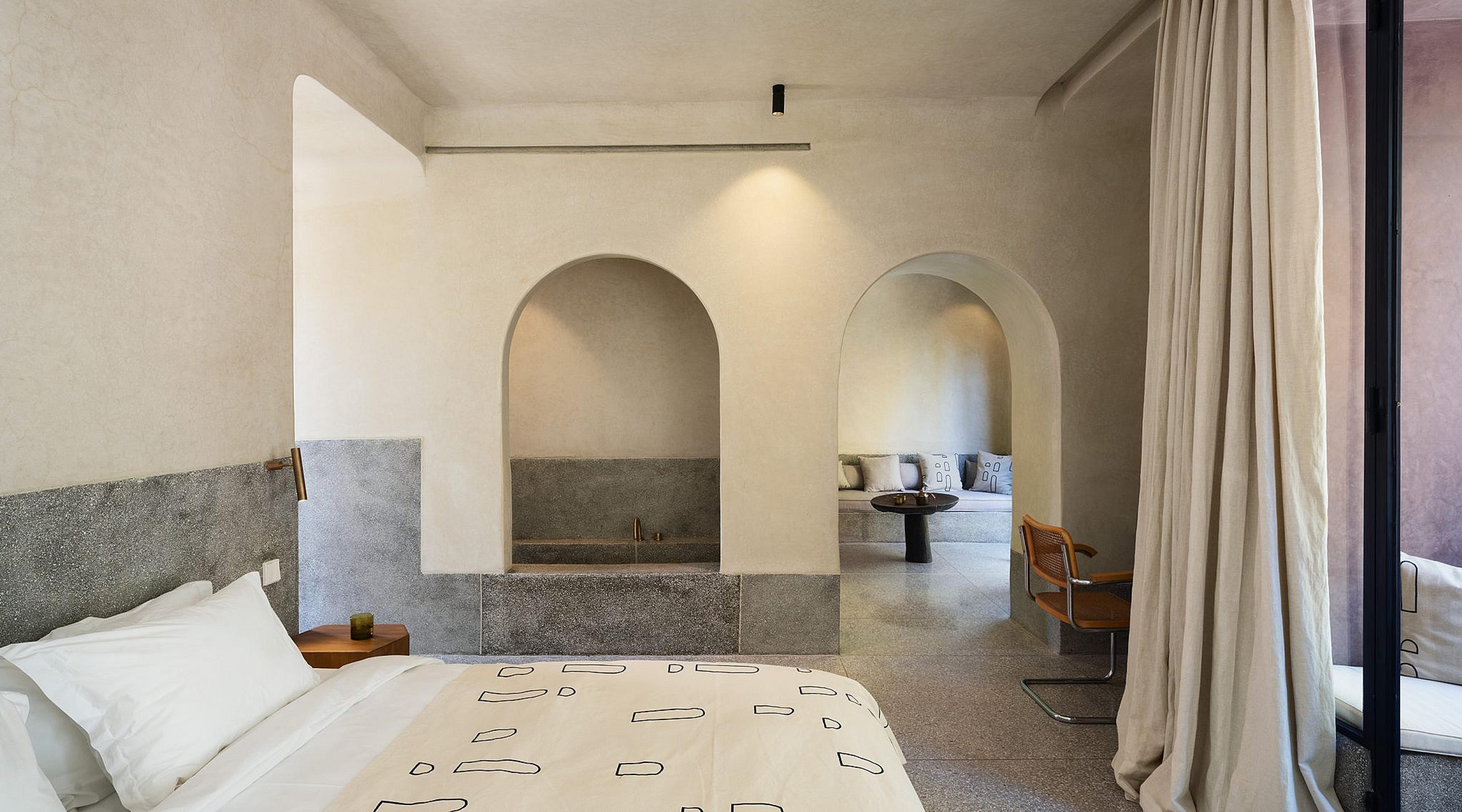 a bedroom with a grey-trimmed bed facing an arched alcove with a sunken bath, and a further archway opening onto a seating area