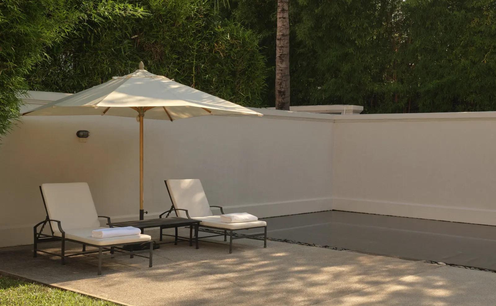 A private pool terrace with two loungers, a white umbrella and folded towels, bordered by a plain wall and trees.