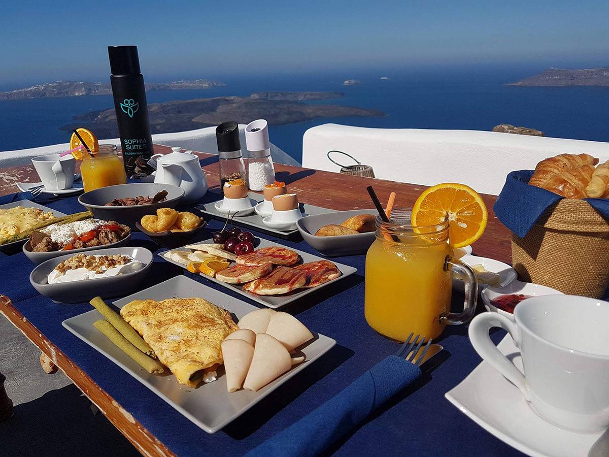 A breakfast table outdoors with an omelette, pancakes, sliced fruit, a jar of orange juice with a slice of orange, coffee cups and a basket of croissants, with the sea and islands behind.