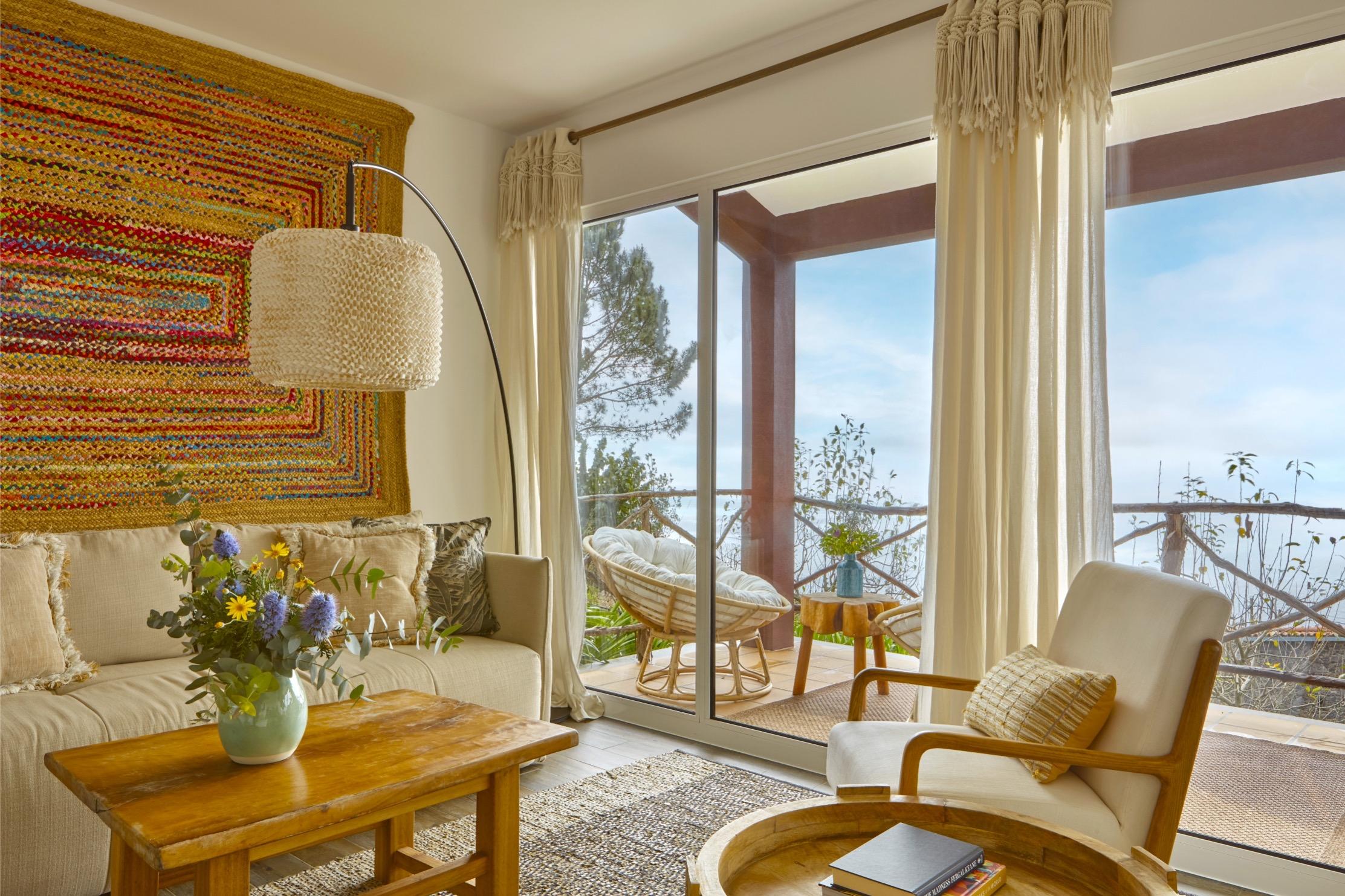 A living room with a cream sofa, a woven-shade arc lamp and a patterned wall hanging, its glass doors open to a balcony with a rattan chair and a view of trees and sky.