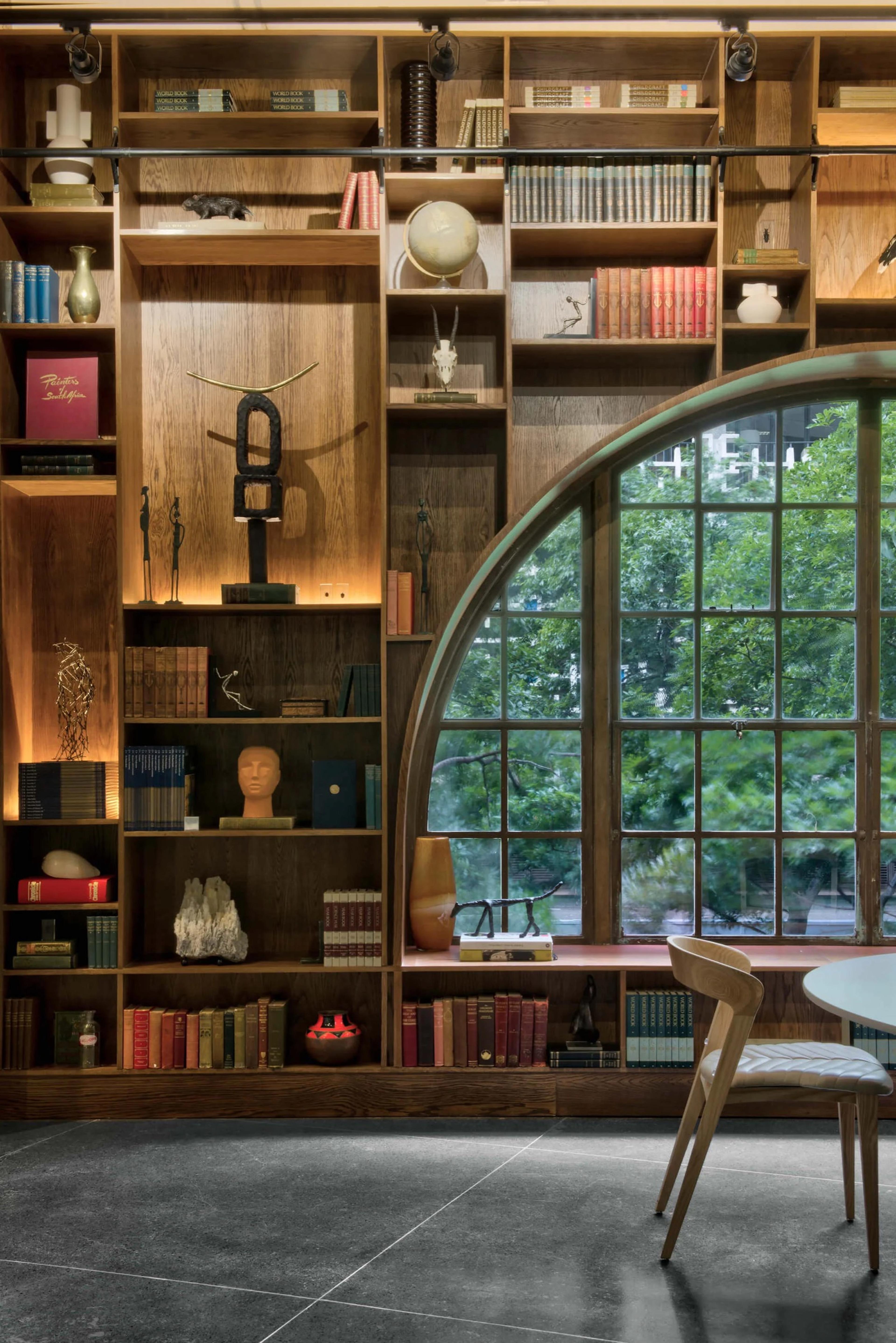 Dark wooden shelves full of books and objects fill the wall beside a round-topped window of small panes, with trees outside and a pale wooden chair at a round table.