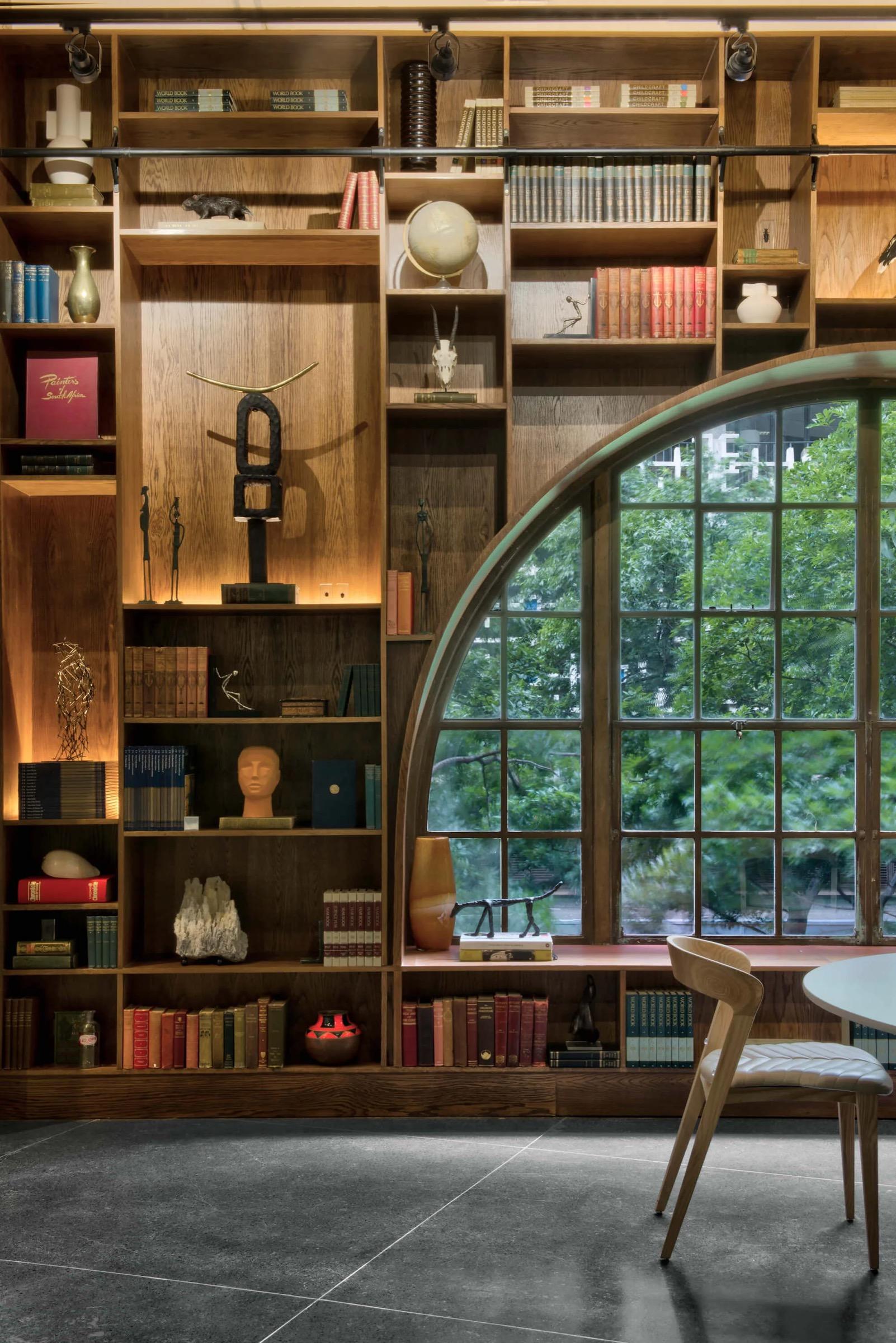Dark wooden shelves full of books and objects fill the wall beside a round-topped window of small panes, with trees outside and a pale wooden chair at a round table.