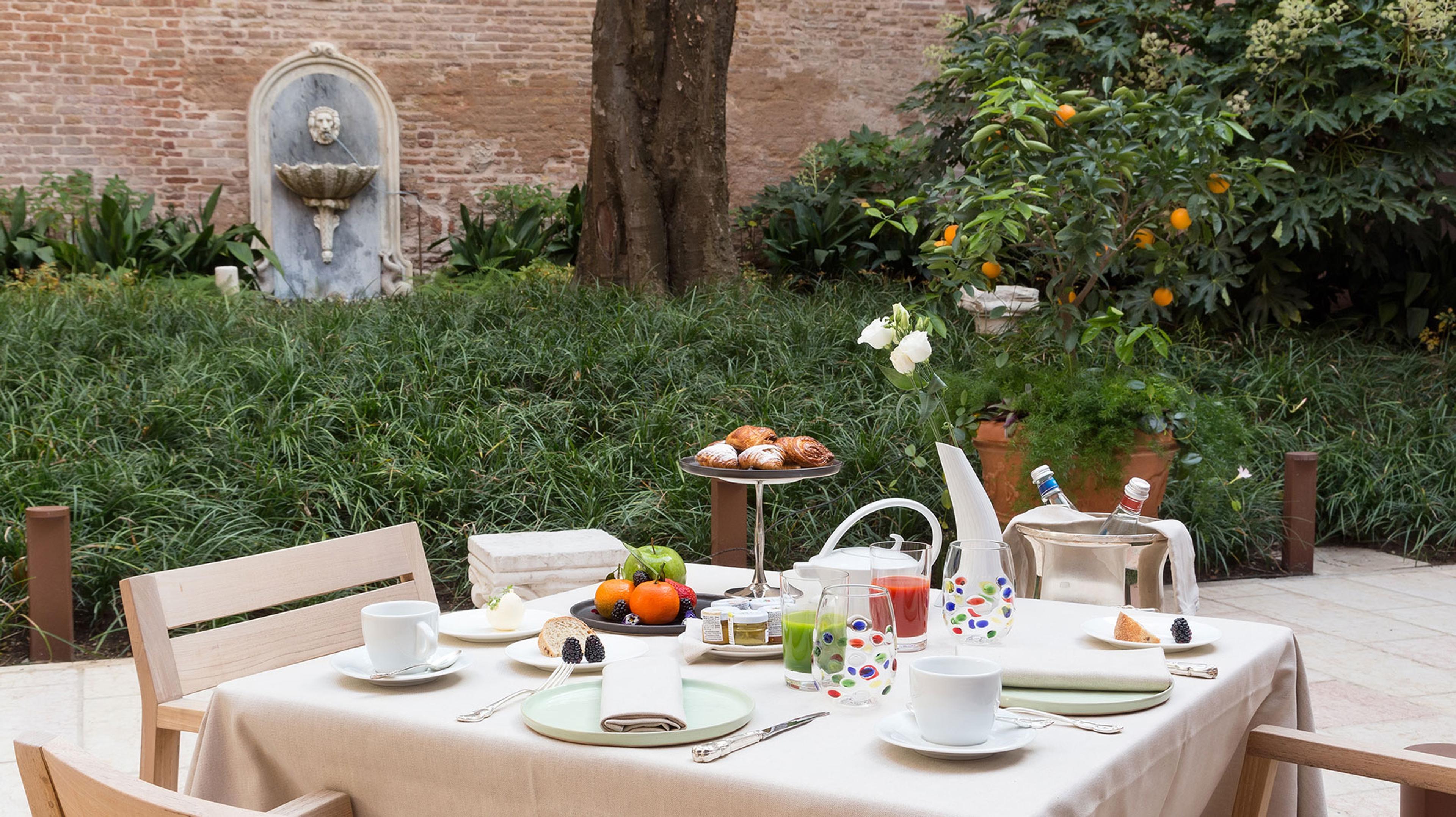 A table laid for breakfast in a garden, with pastries, fruit, juices and coffee, next to a stone wall fountain and an orange tree.