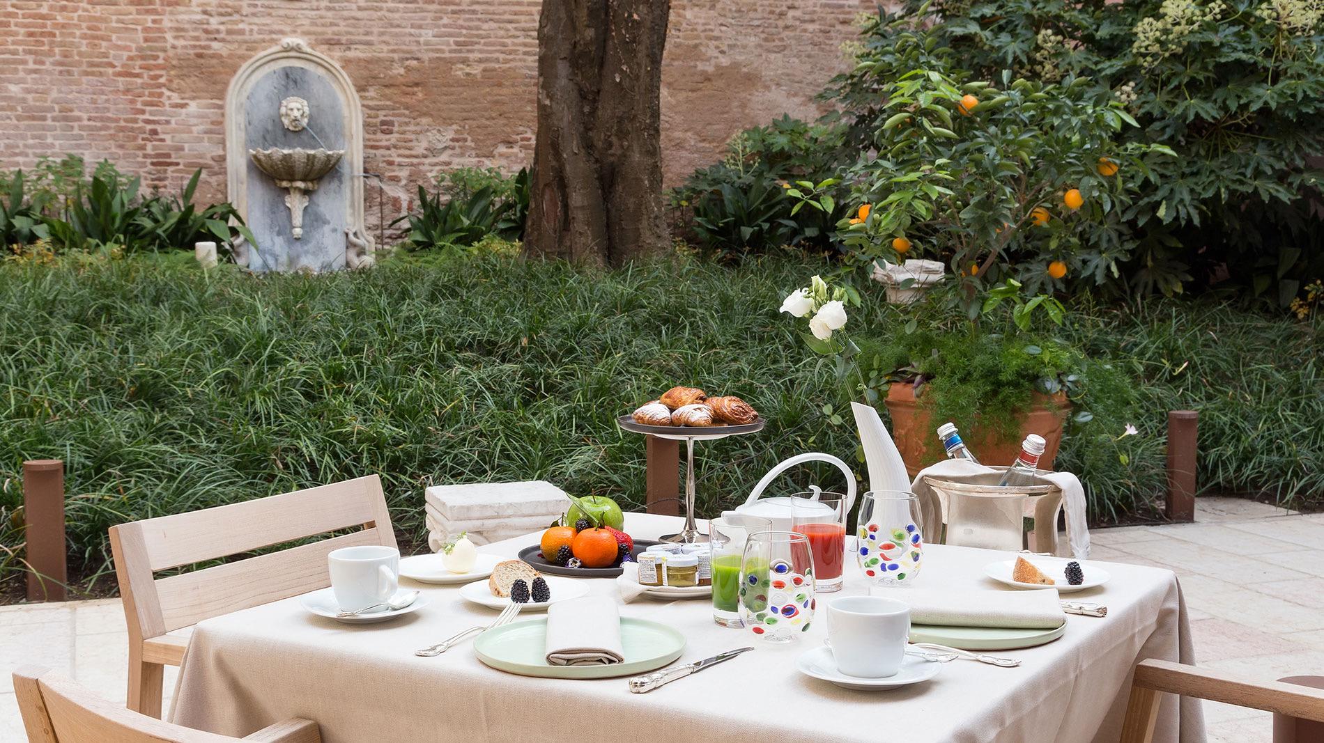 A table laid for breakfast in a garden, with pastries, fruit, juices and coffee, next to a stone wall fountain and an orange tree.