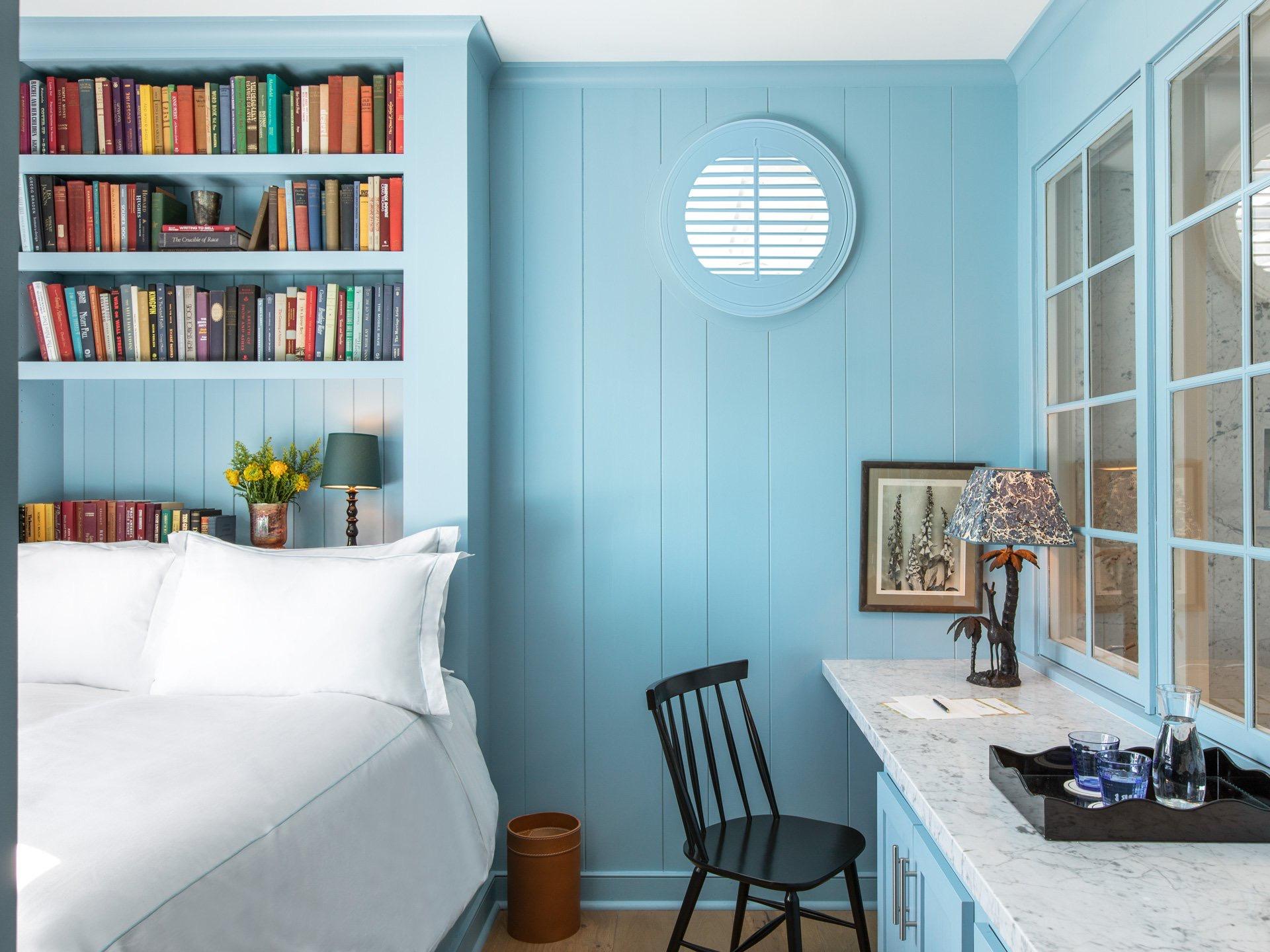 A pale blue bedroom with a built-in bookcase of colour-ordered books, a round shuttered window, and a marble desk with a carafe and glasses.
