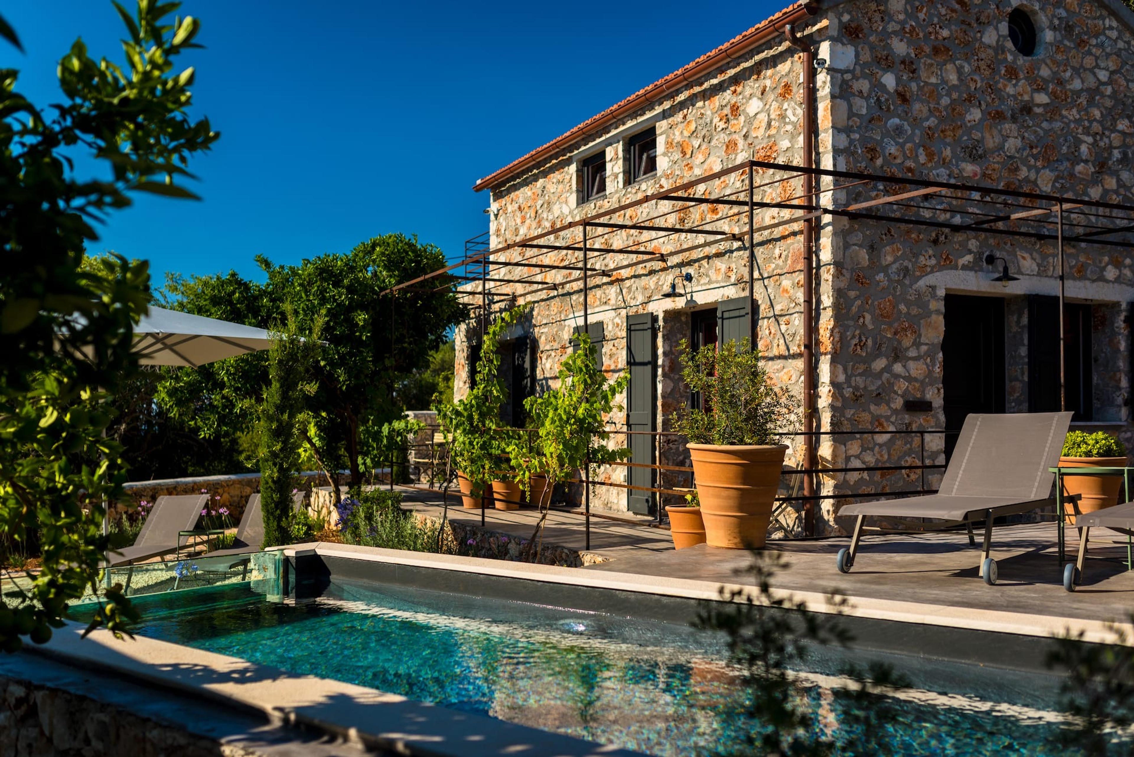A two-storey stone house with a terracotta roof, seen across a plunge pool with sun loungers, a shade umbrella and terracotta pots of plants on the terrace.