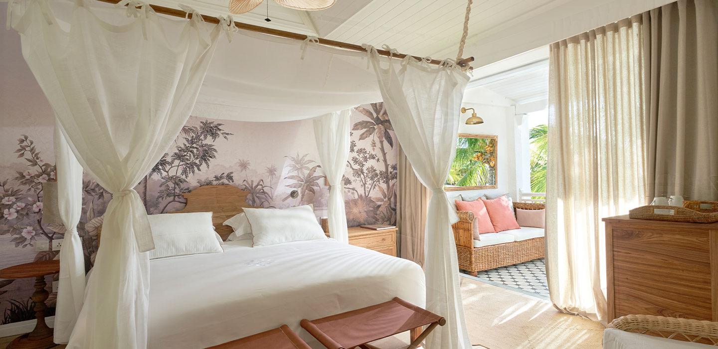 A four poster bed with sheer white curtains stands against a mural of tropical leaves, with a cushioned seating nook and a wooden chest beyond it.