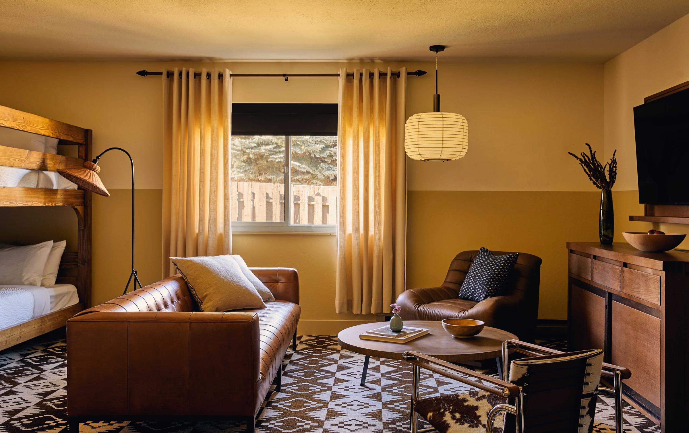 A hotel living room with a tufted brown leather sofa and armchair, a wooden bunk bed, a patterned rug and curtained window.