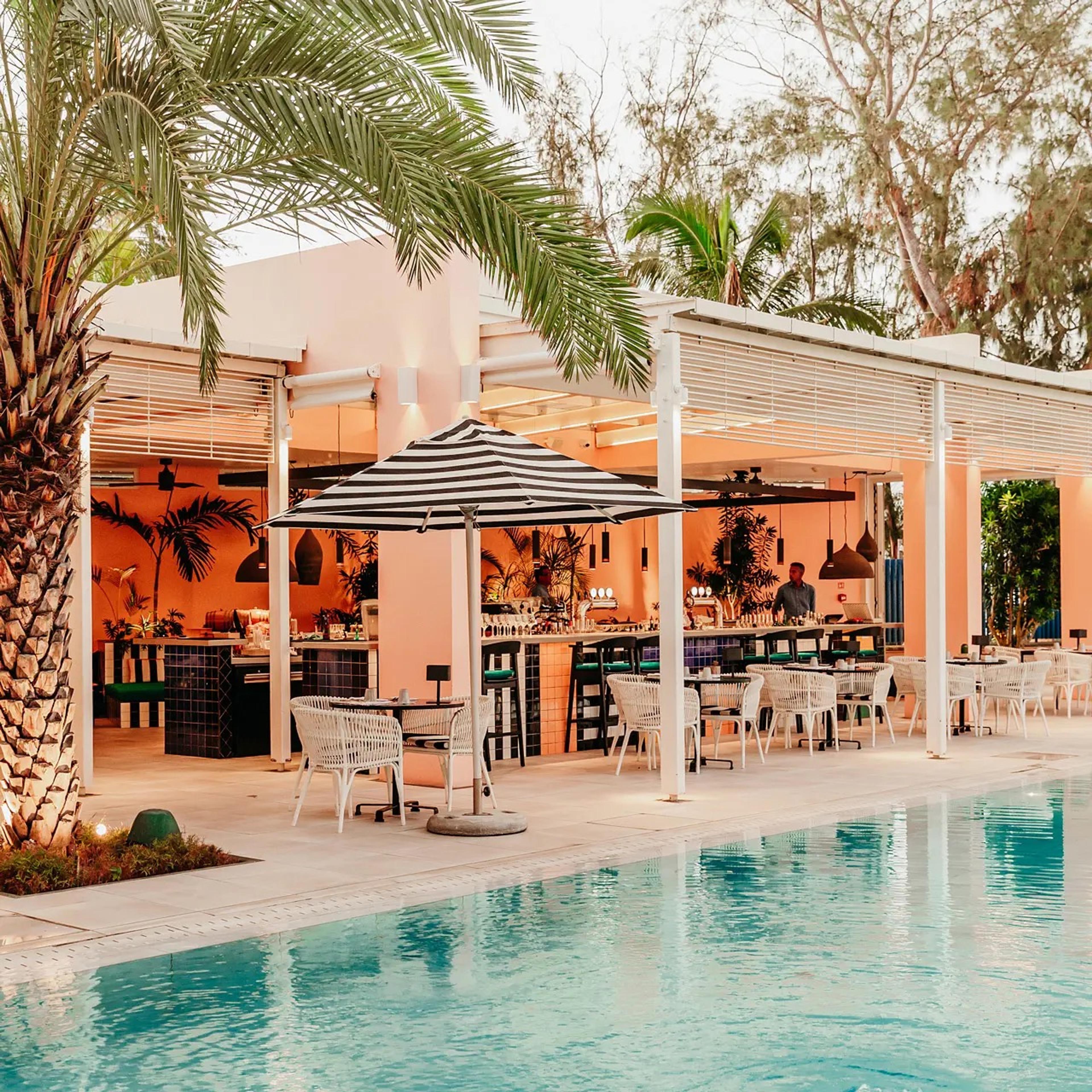 A poolside bar and dining area shaded by a striped umbrella and a palm tree, seen across the pool at dusk.