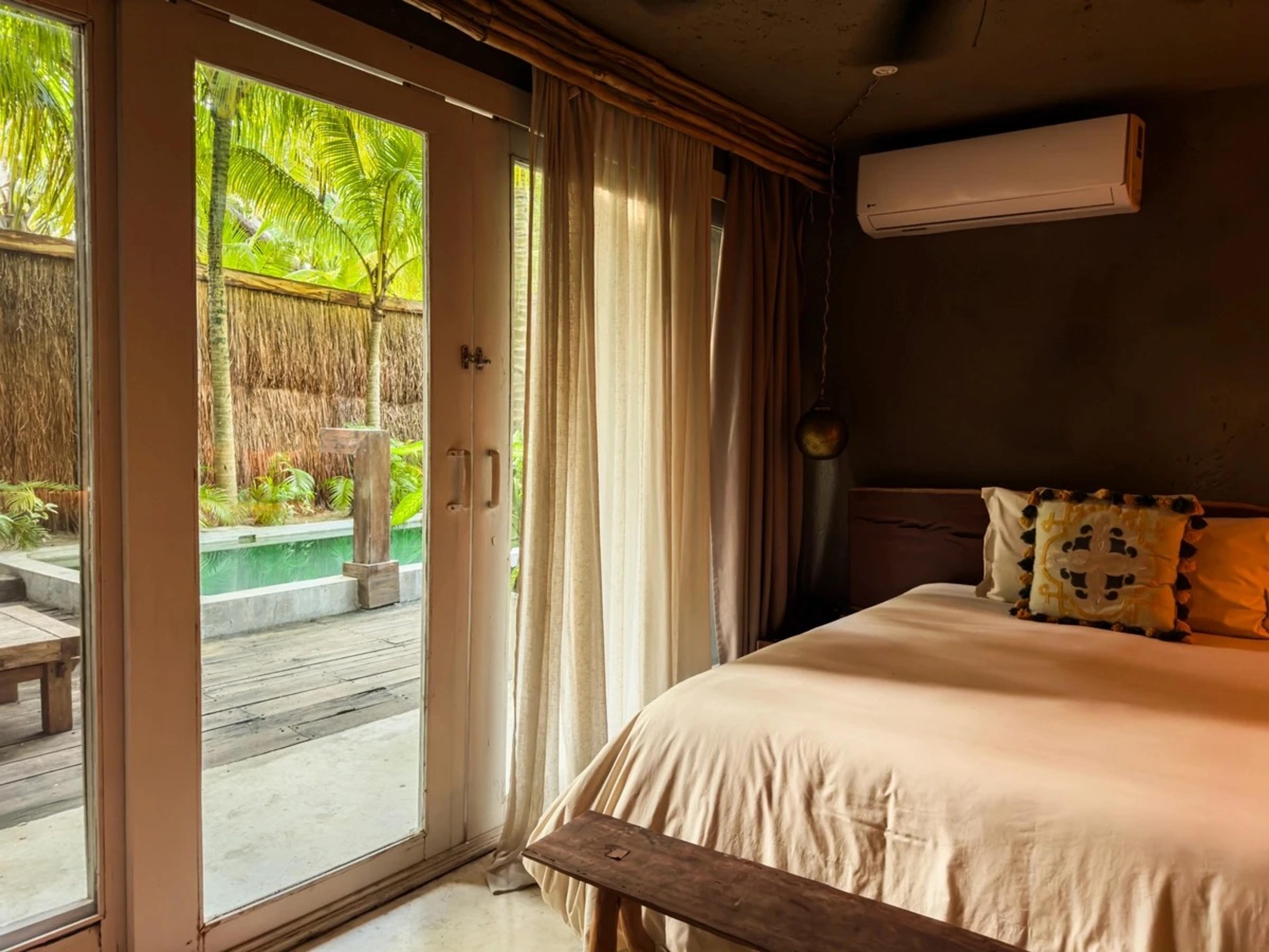 A bedroom with open glass doors leading onto a pool deck surrounded by palm trees.