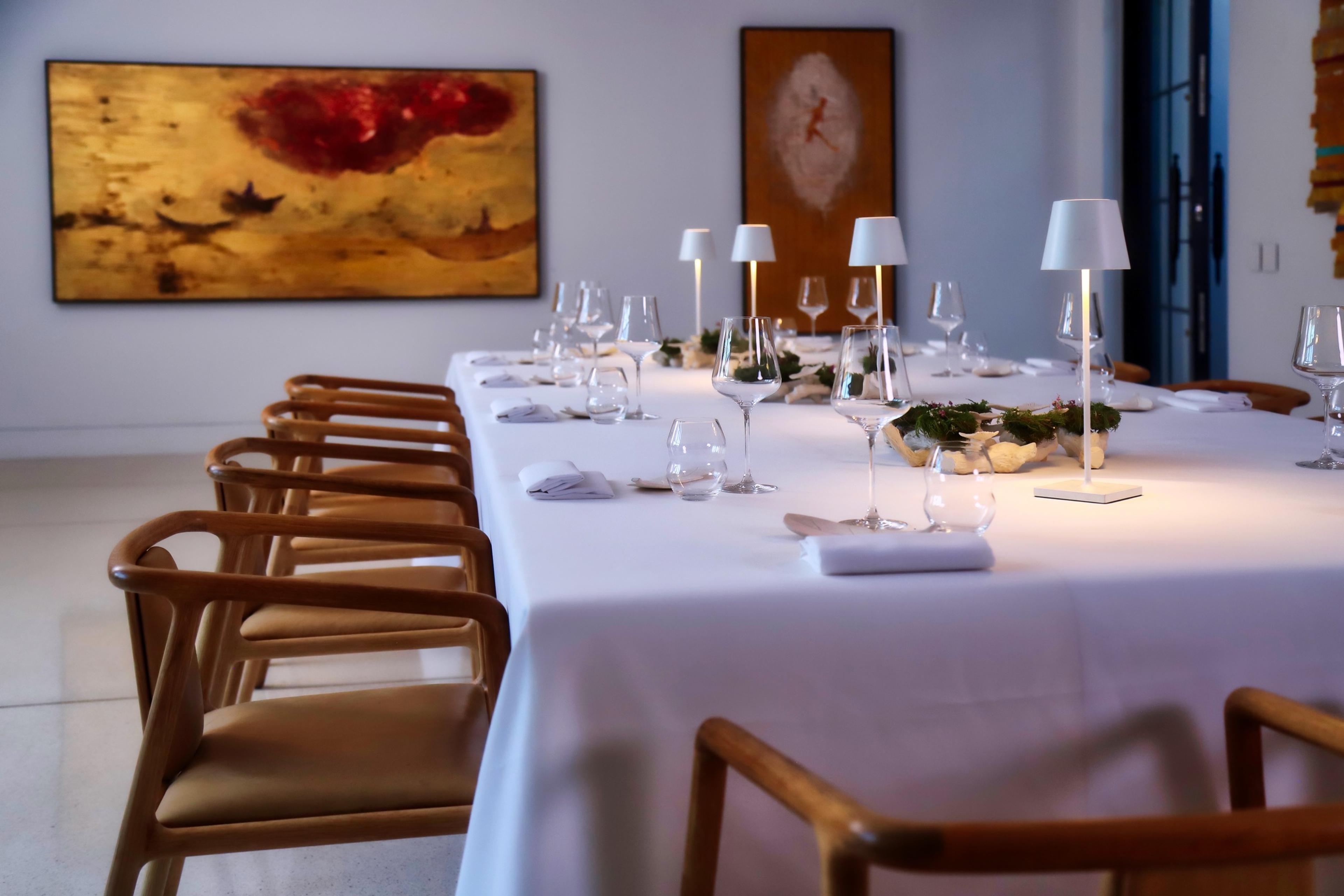 A long table laid with a white cloth, glasses and small lamps, in a dining room hung with paintings