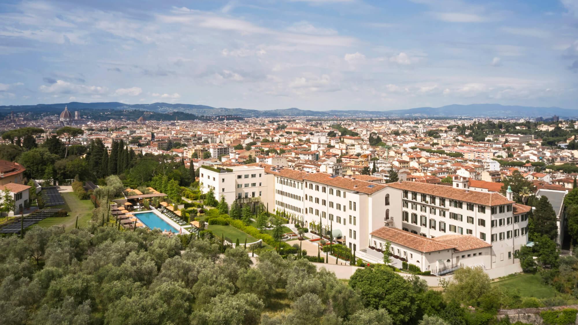 an aerial view of a cream colored villa complex with a swimming pool and gardens, overlooking Florence's rooftops and the Duomo in the distance