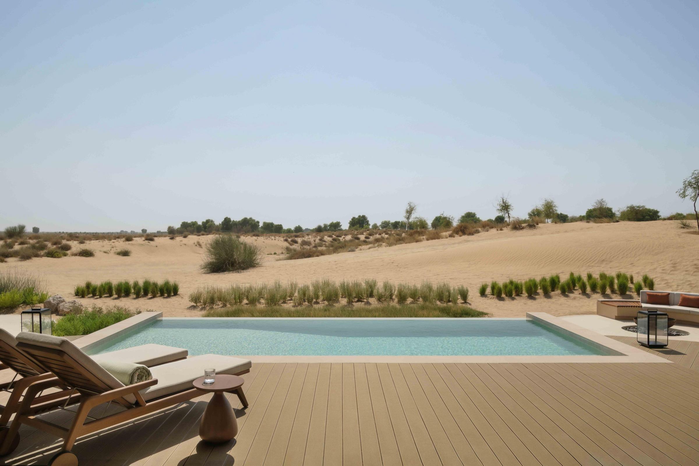 A rectangular pool on a wooden deck with two loungers, bordered by desert scrub and sand dunes under a clear sky.
