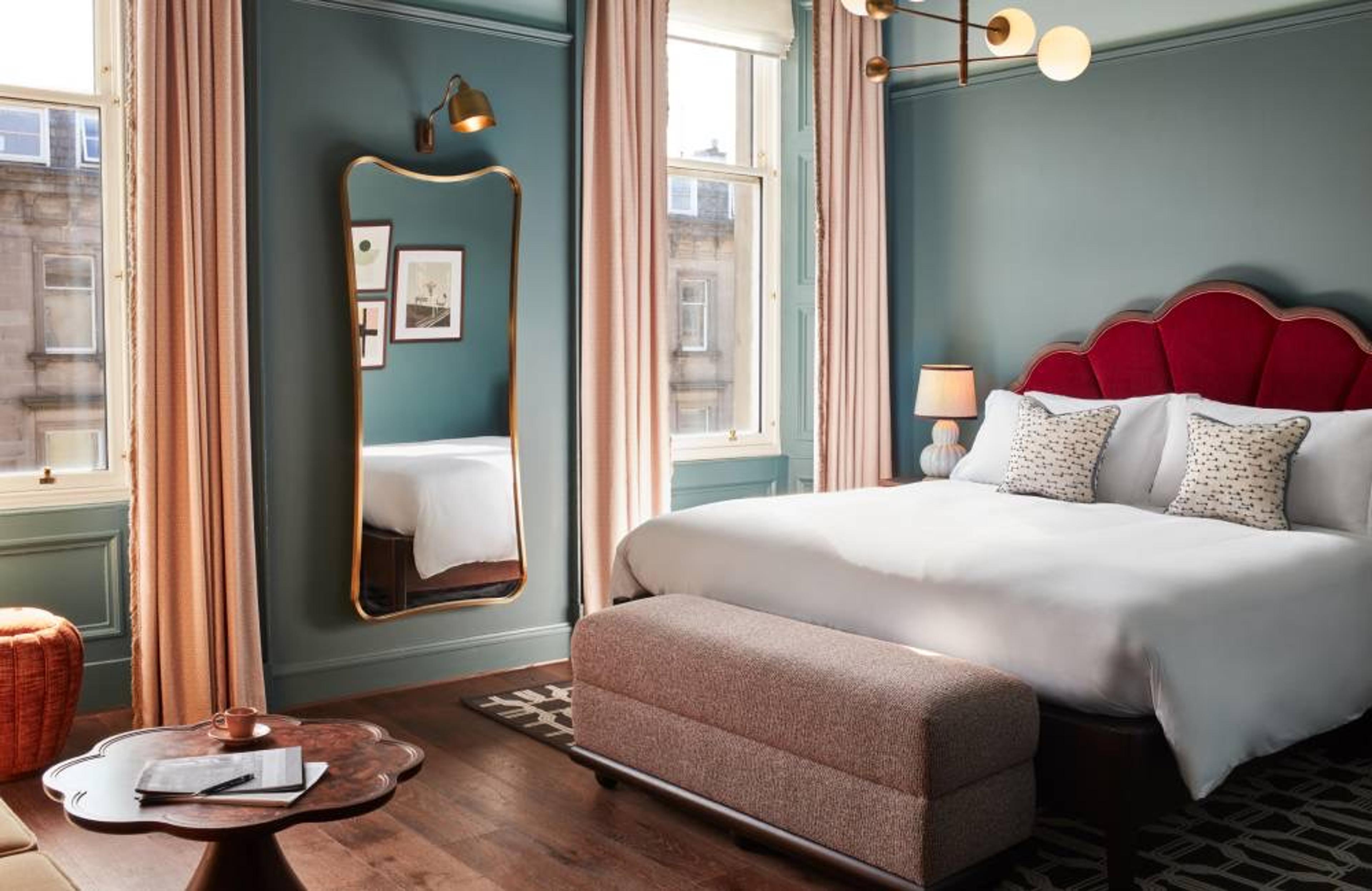 A larger bedroom with tall windows, a red scalloped headboard and an upholstered bench at the foot of the bed.