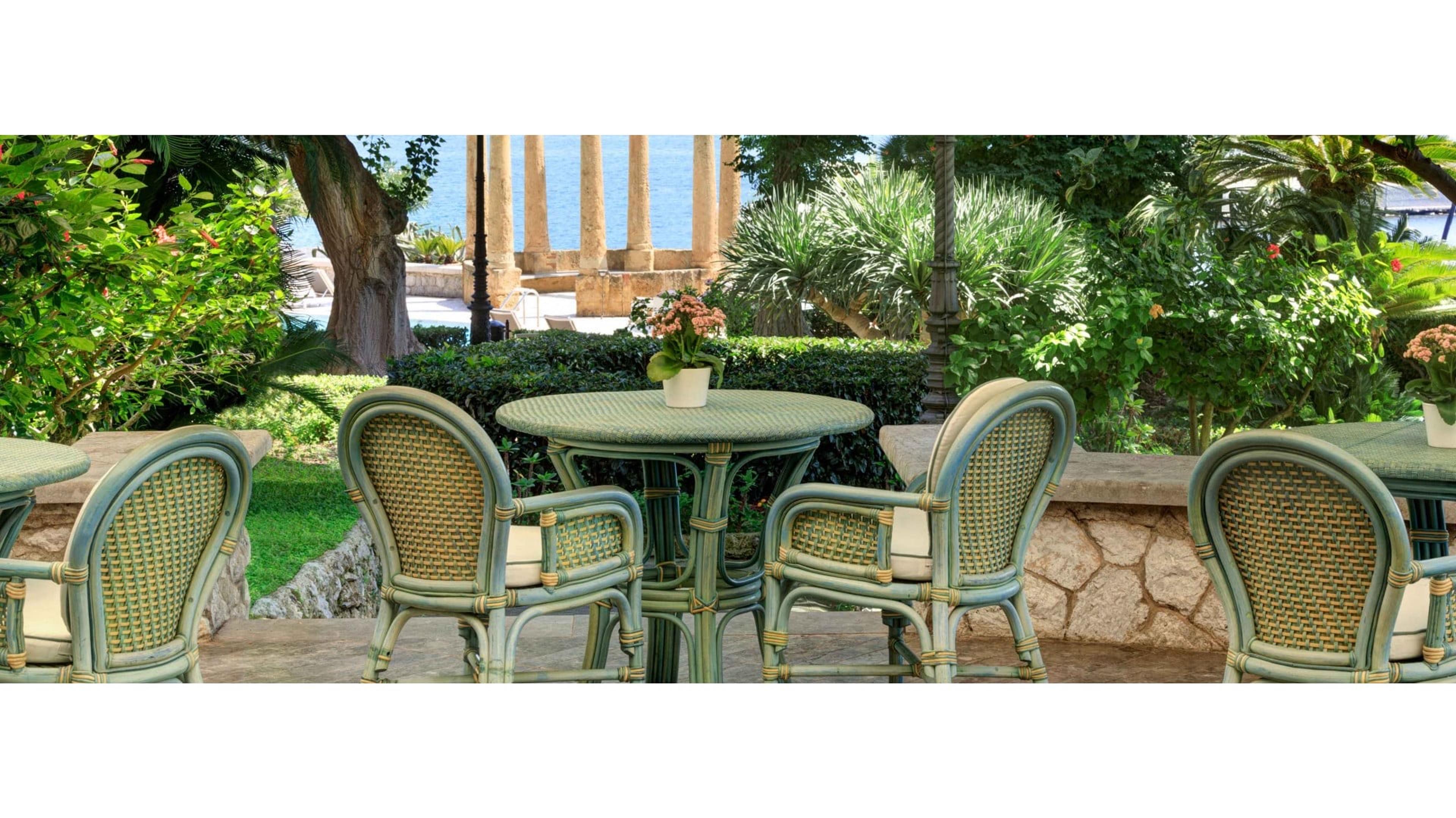 Wicker chairs around small tables on a stone terrace, looking past a hedge and the ruins of a classical colonnade toward the sea.