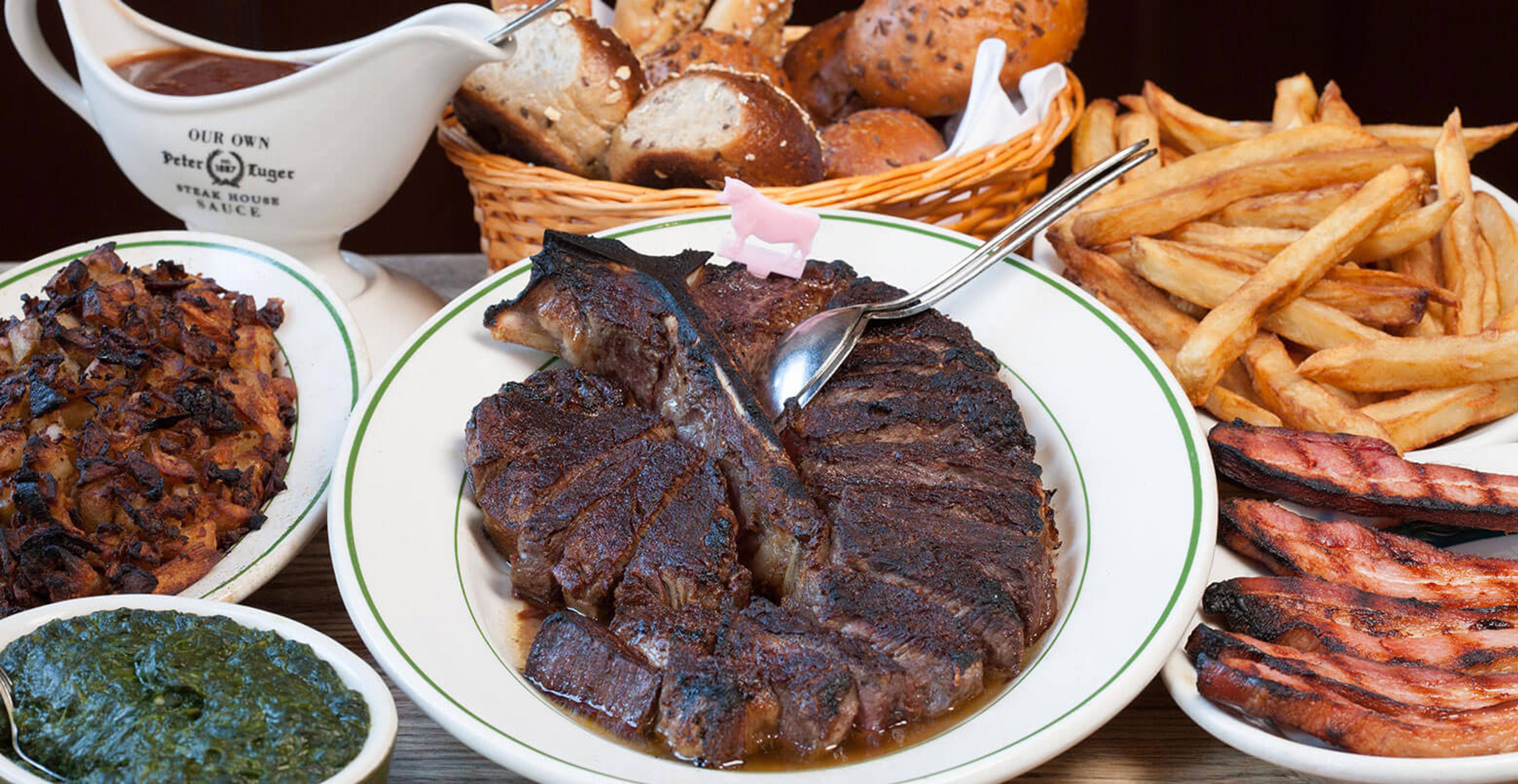 A sliced porterhouse steak on a white platter, with fries, creamed spinach and a bread basket around it